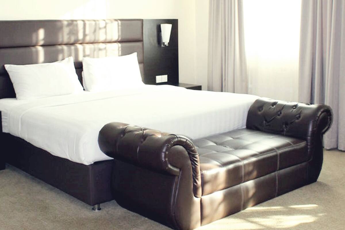 A hotel room with a large bed, a dark brown leather sofa, a nightstand, and a window with curtains.