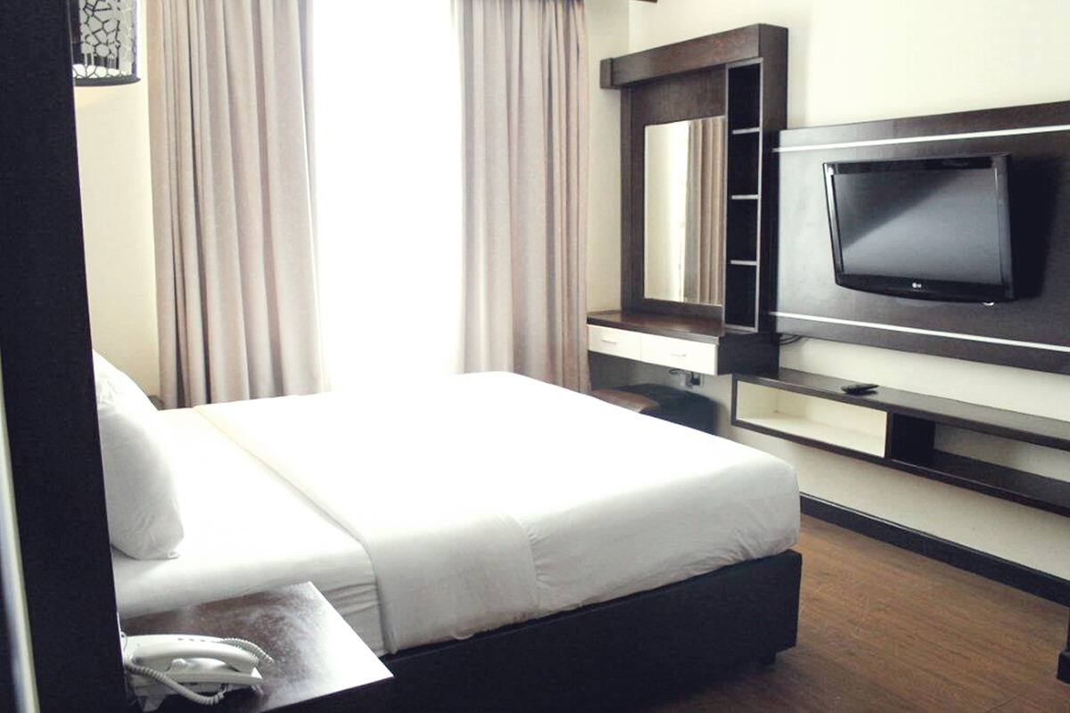 A hotel room with a bed, a TV mounted on the wall, a bedside table with a phone, and a window with curtains.