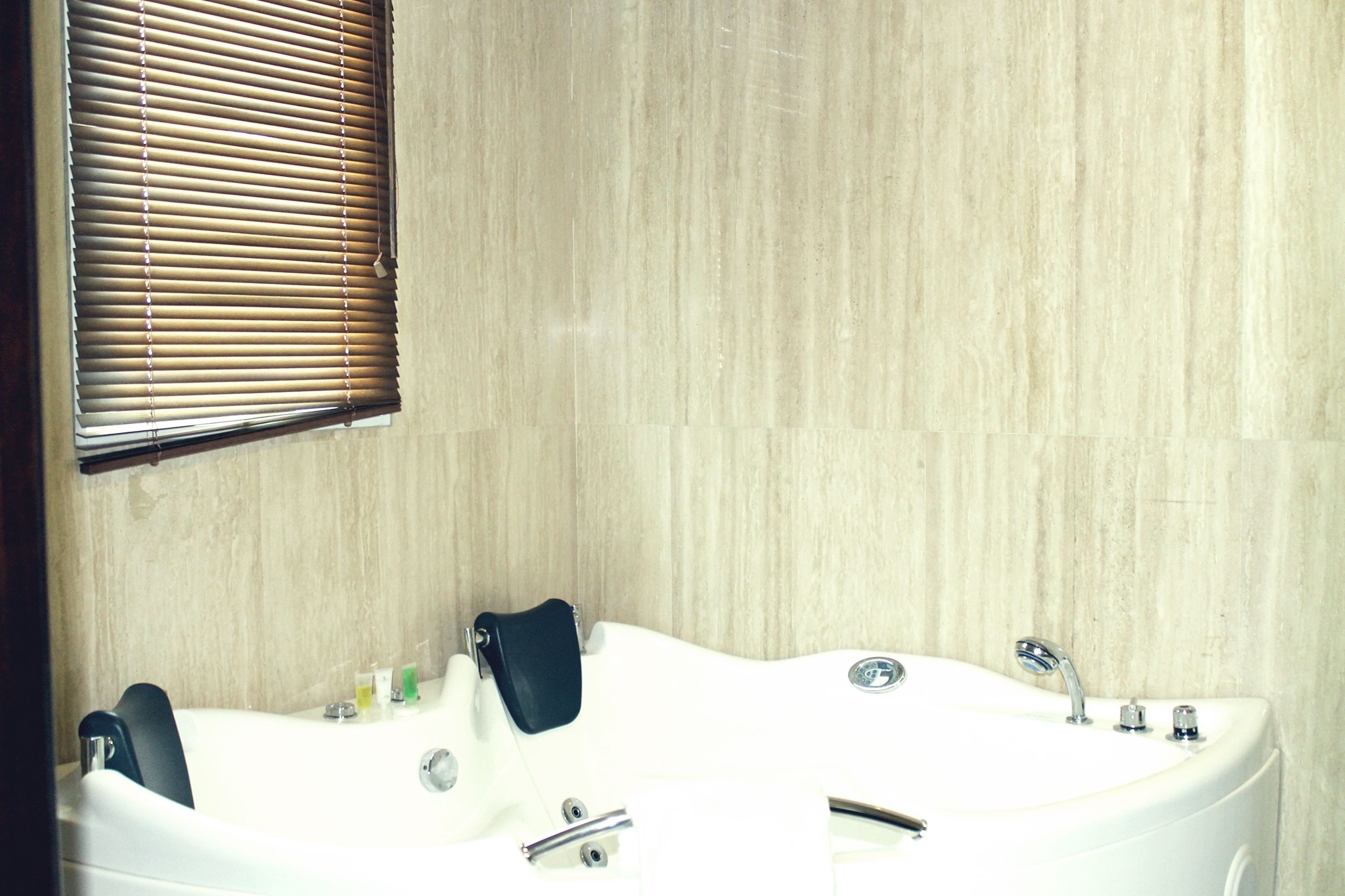 A bathroom with a large bathtub, a window with blinds, and a tiled wall.
