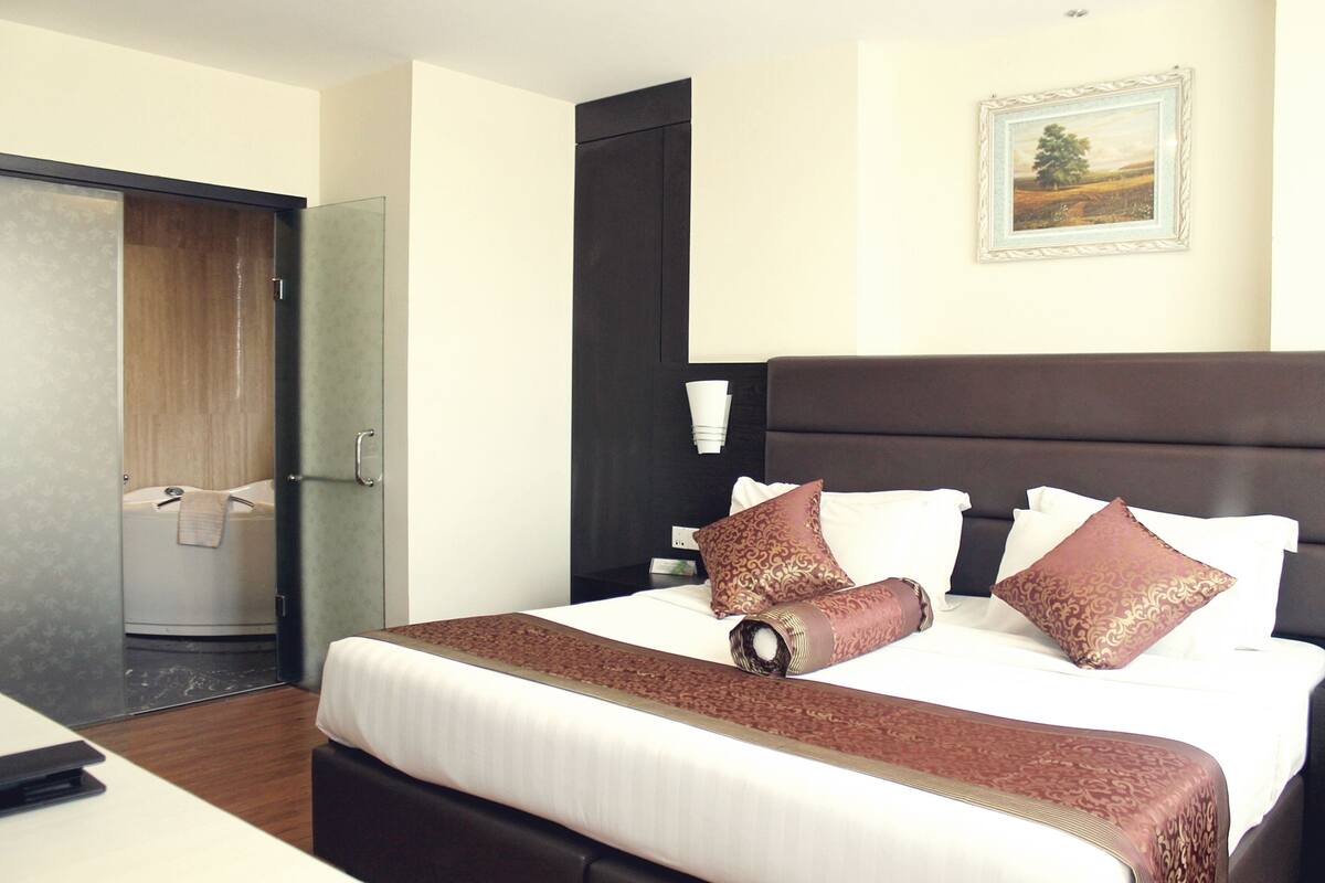 A hotel room with a large bed, a dark headboard, a framed picture on the wall, and a glass door leading to a bathroom.