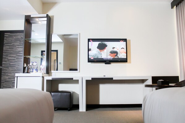 A hotel room with a TV mounted on the wall, a desk with a mirror, and a bed with a headboard. - The BCC Hotel & Residence (Batam)