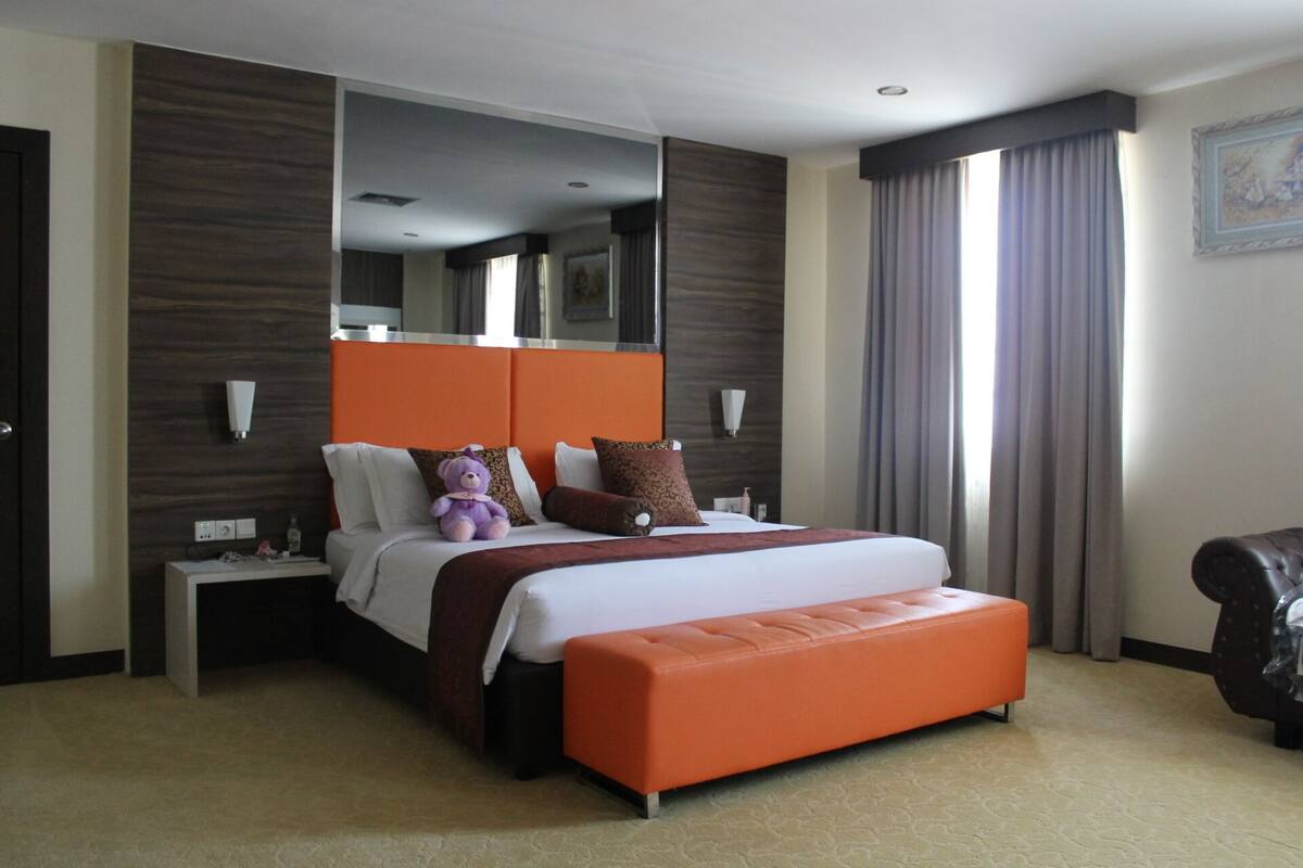 A hotel room with a large bed, an orange headboard, a nightstand, a mirror, a window with curtains, and a chair.