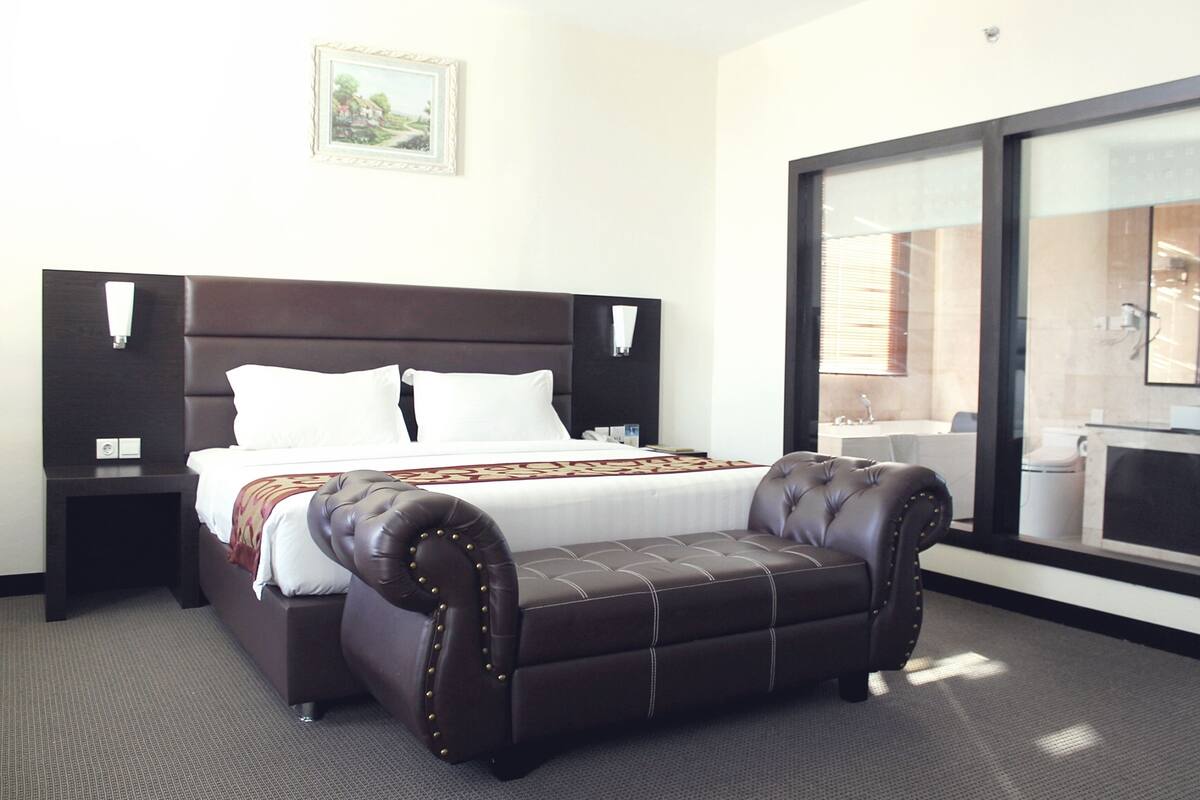 A hotel room with a large bed, a leather ottoman, a nightstand, a wall-mounted lamp, and a window with a view of a bathroom.