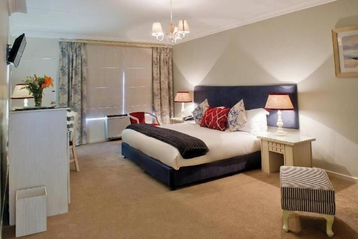 Una habitación de hotel con cama, lámparas de noche, una silla, una mesita y televisor.