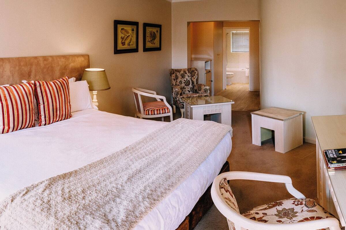 Habitación de hotel con una cama grande, una mesita de noche con lámpara, una silla, una pequeña mesa y una revista sobre la mesa.