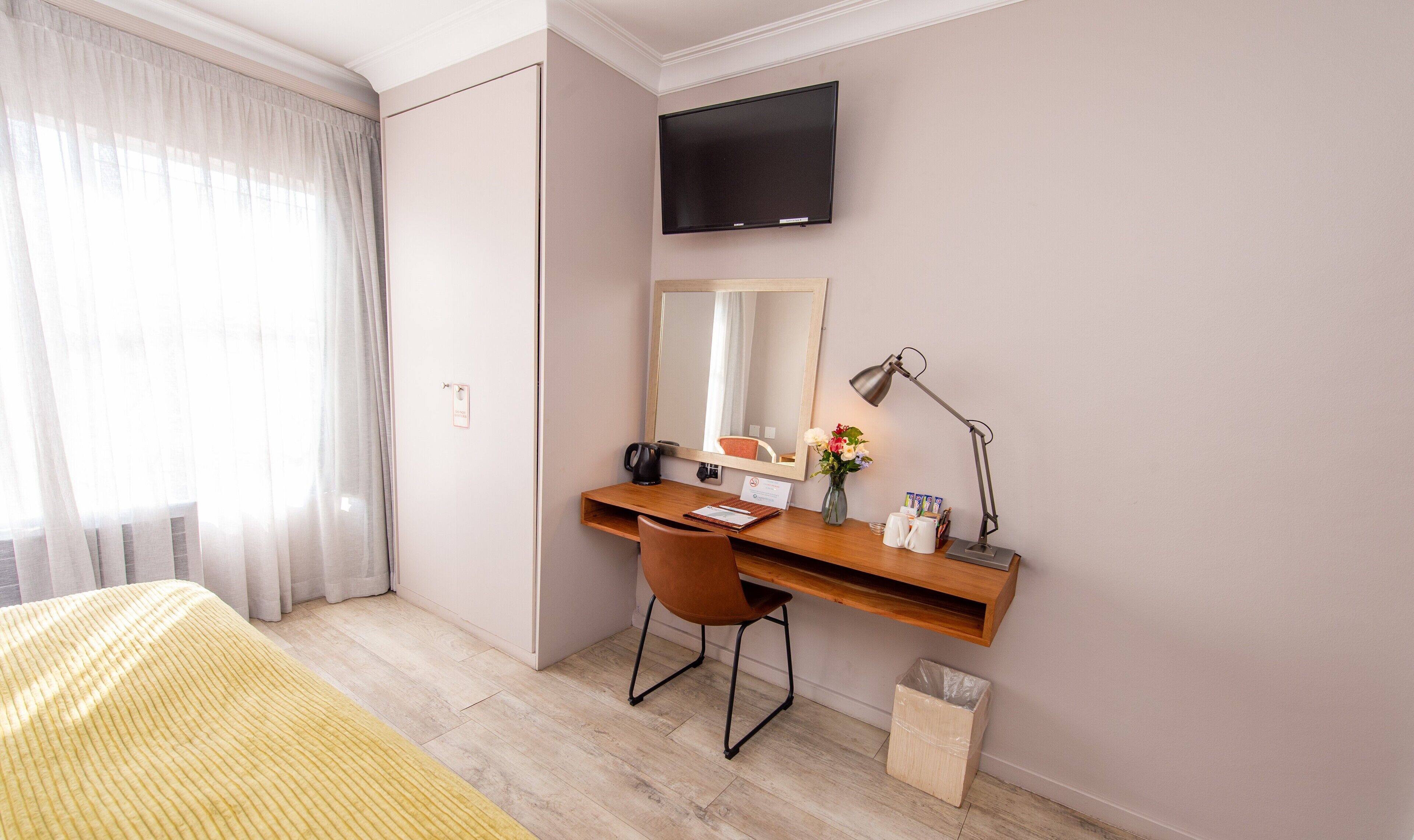 A hotel room with a bed, a desk with a chair, a flat-screen TV, a mirror, a desk lamp, a trash bin, and a window with curtains.
