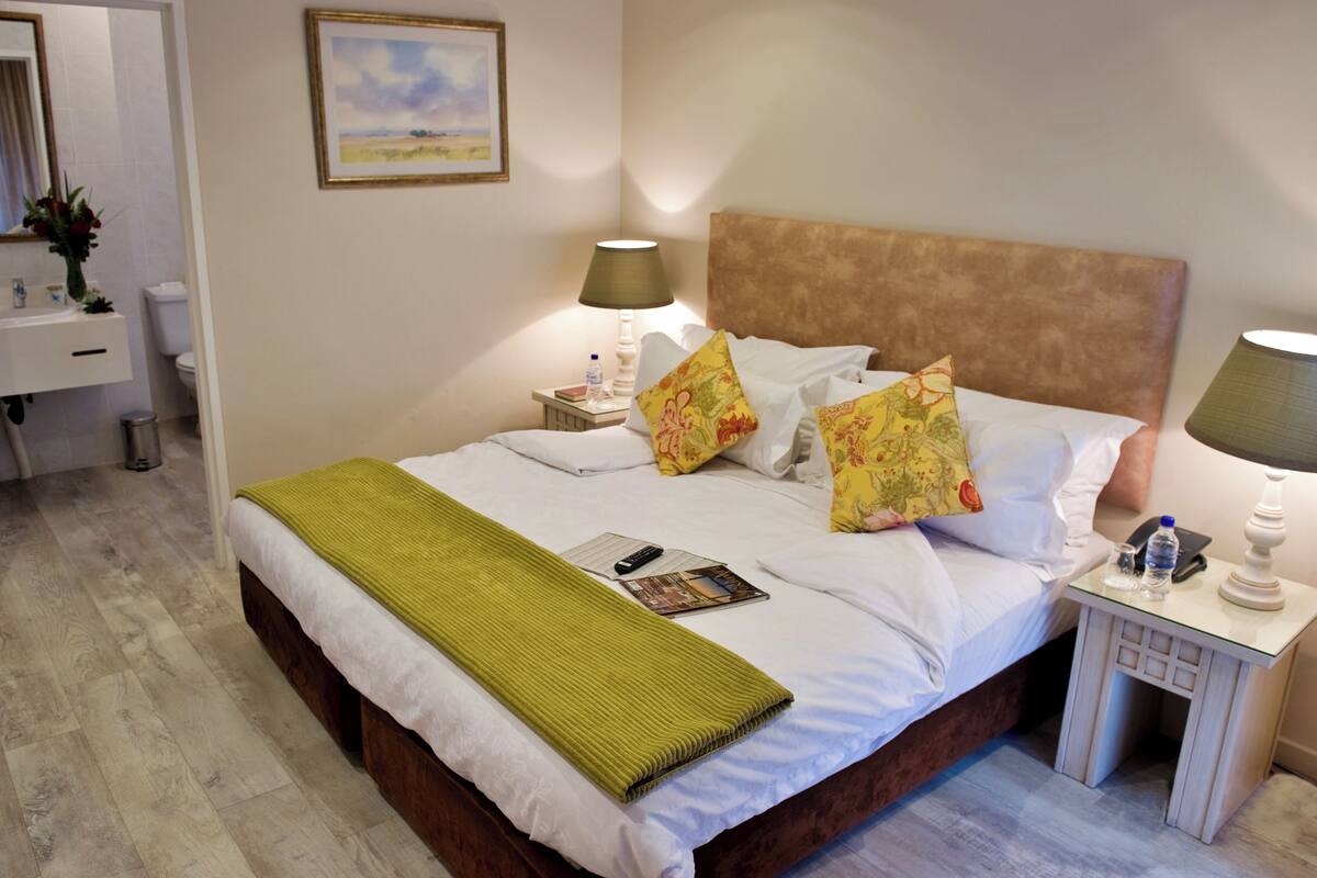 Una cama bien hecha con un plaid verde, dos lámparas de noche y una mesita de noche con una botella de agua y un teléfono.