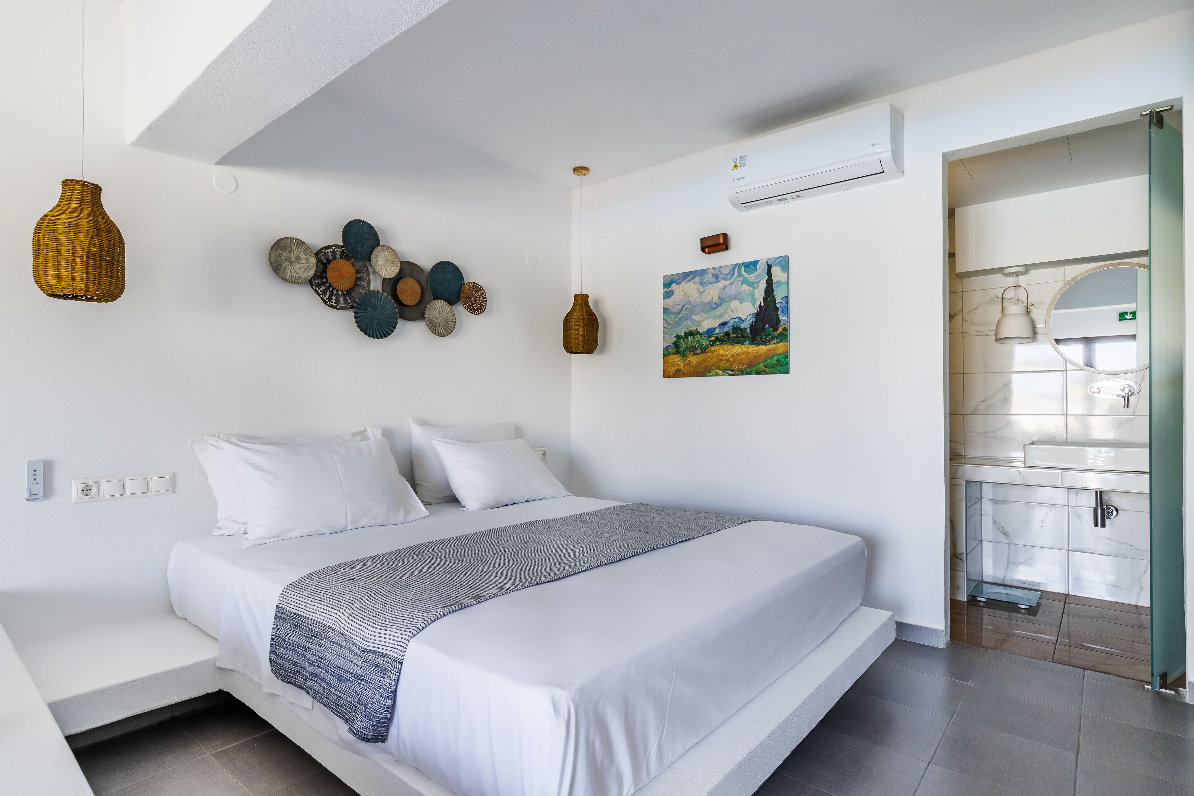 Un dormitorio moderno con una cama grande, una mesita de noche, aire acondicionado de pared y un baño visible a través de una puerta abierta.