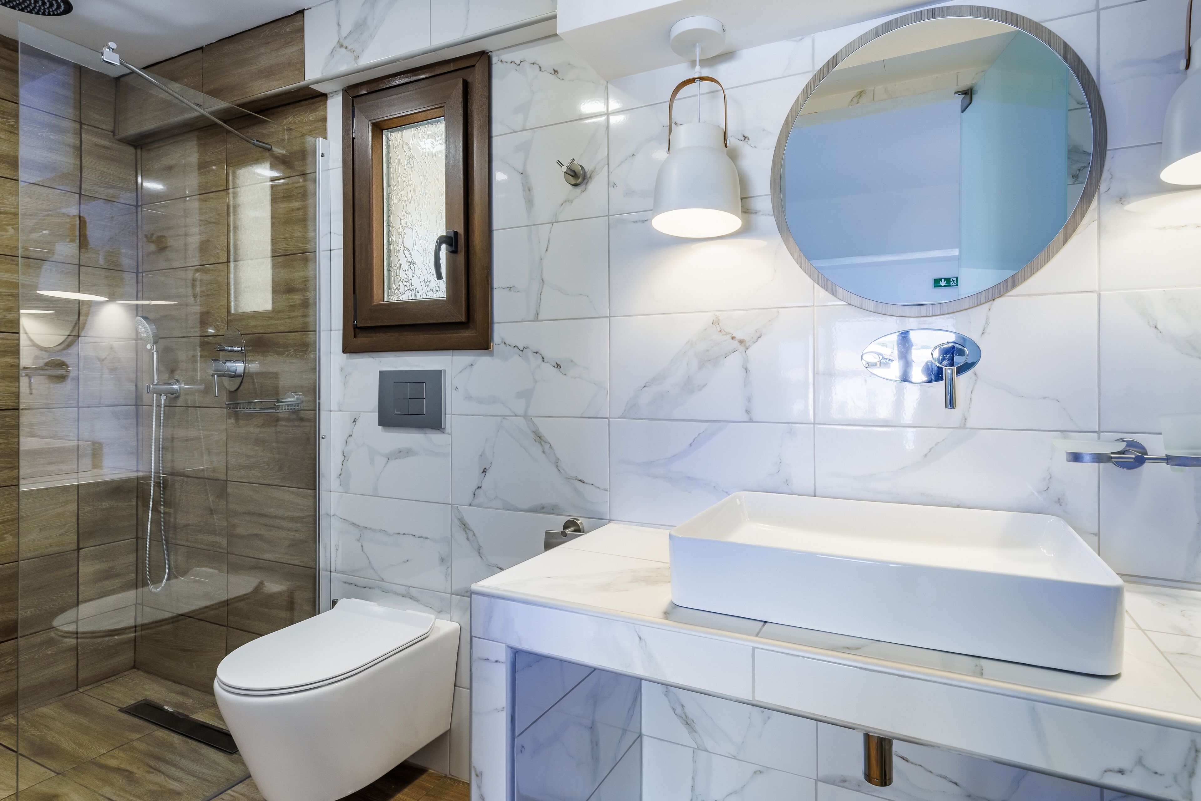 Ein modernes Badezimmer mit einer Glaskabine zur Dusche, einer weißen Toilette, einem großen ovalen Spiegel und einem weißen Waschbecken mit Marmorplatte.