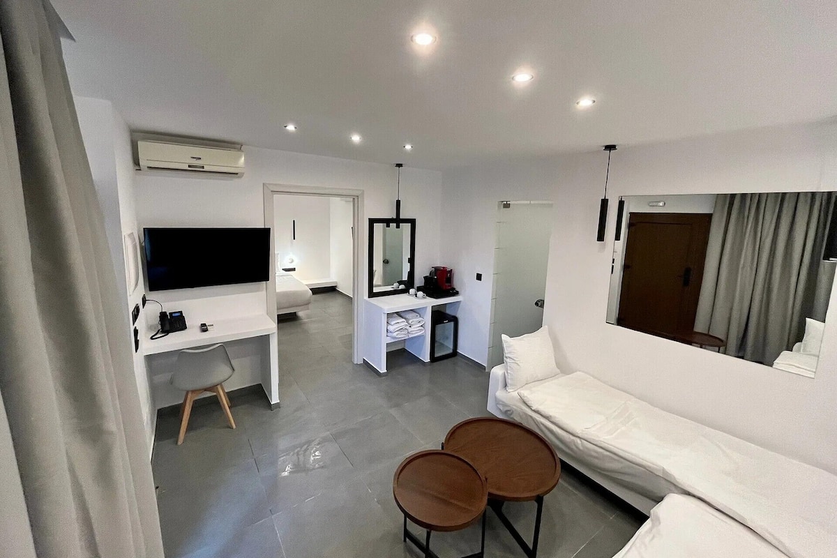 A modern hotel room with a flat-screen TV, a desk with a mirror, a sofa, and two small round tables.