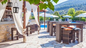 Uma área de bar ao ar livre com móveis de vime, piscina e montanhas ao fundo.