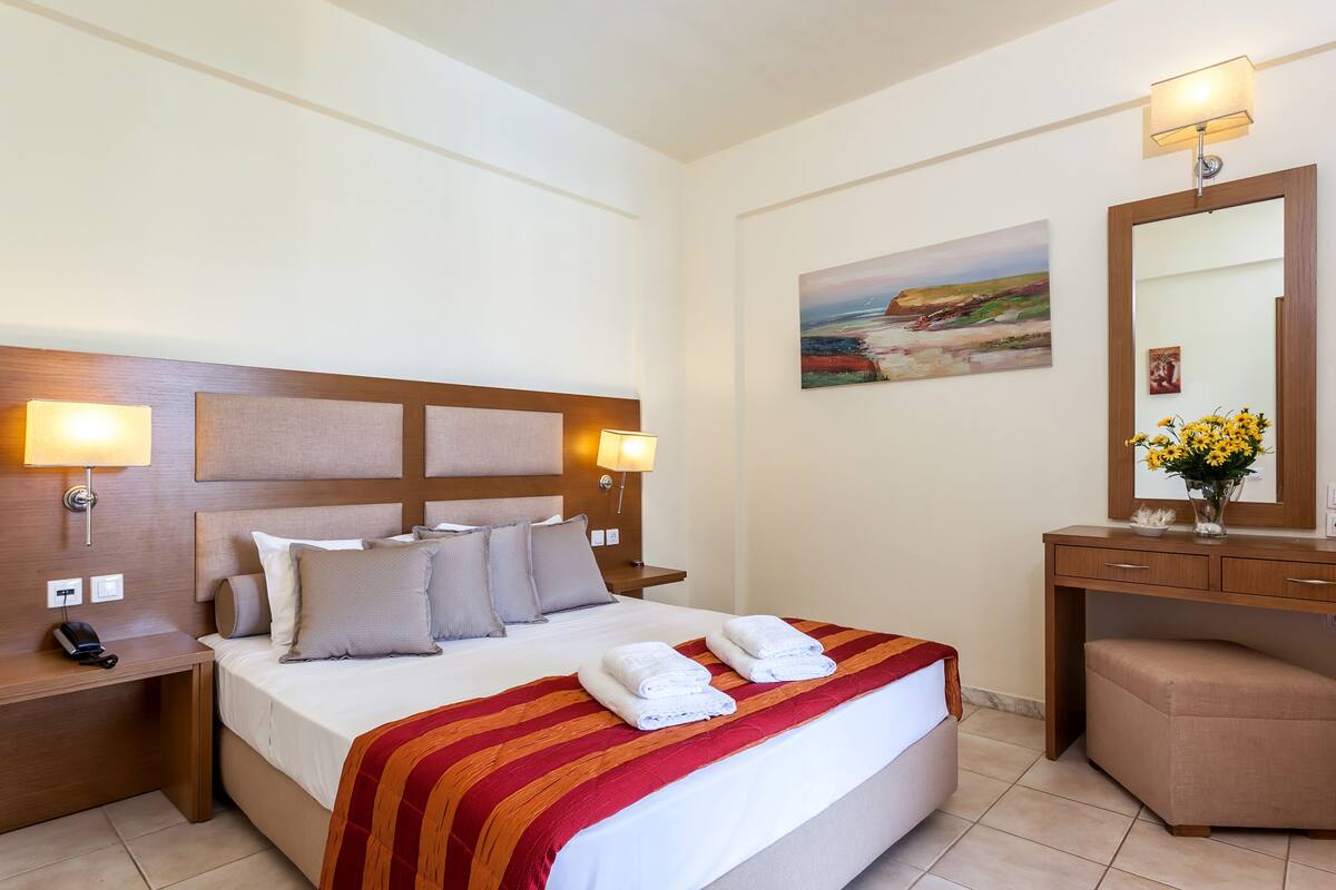 Una habitación de hotel con una cama grande, dos lámparas de noche, una mesita de noche con un teléfono, un escritorio con un espejo y un cuadro en la pared.