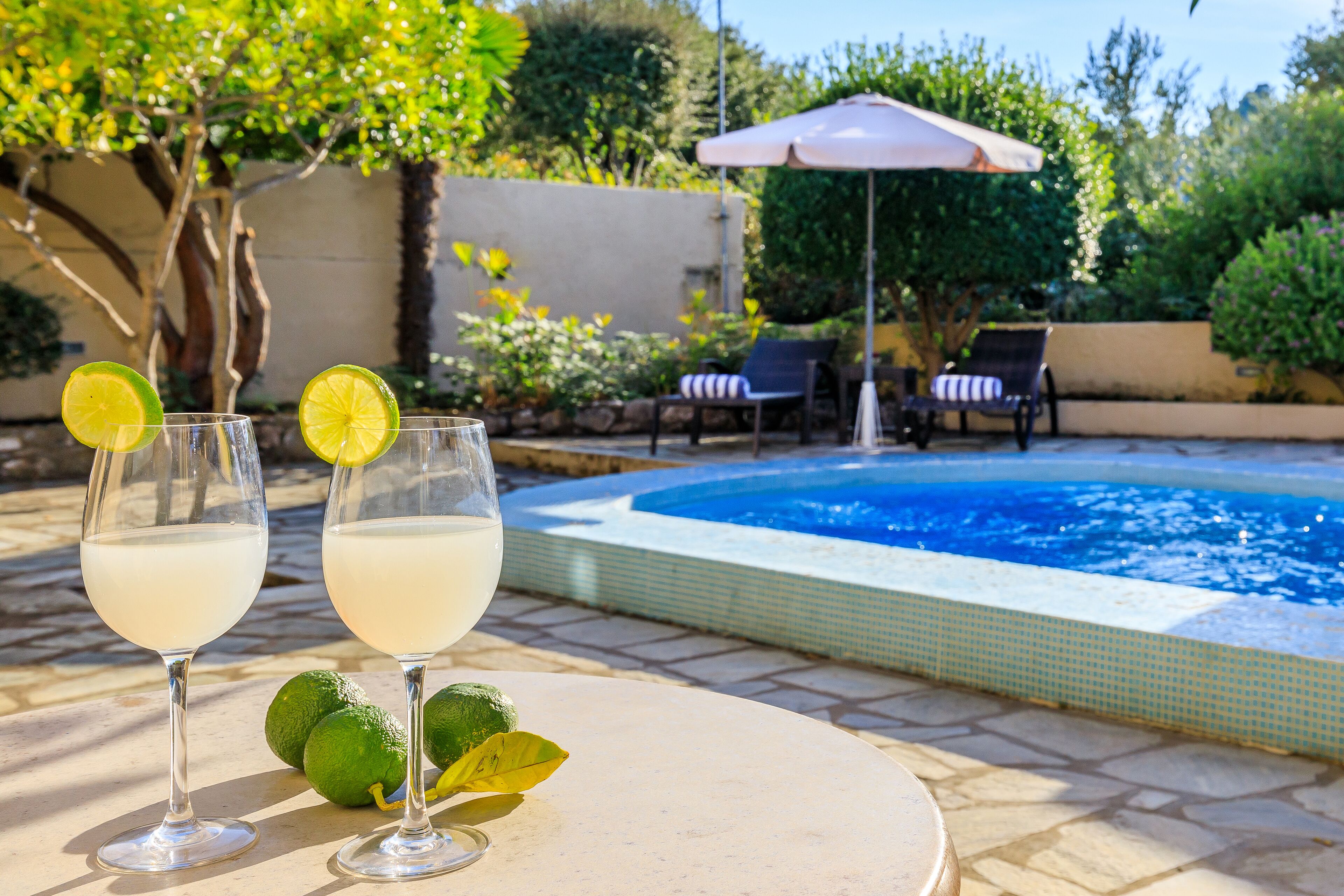 Two glasses of a light-colored beverage with lime slices on a patio table, a swimming pool, and outdoor seating with umbrellas.