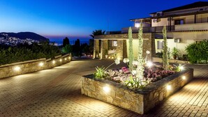 Uma área externa bem iluminada, com um vaso de pedra cheio de flores e plantas, um caminho pavimentado e um edifício com várias varandas e vista para a cidade ao entardecer.