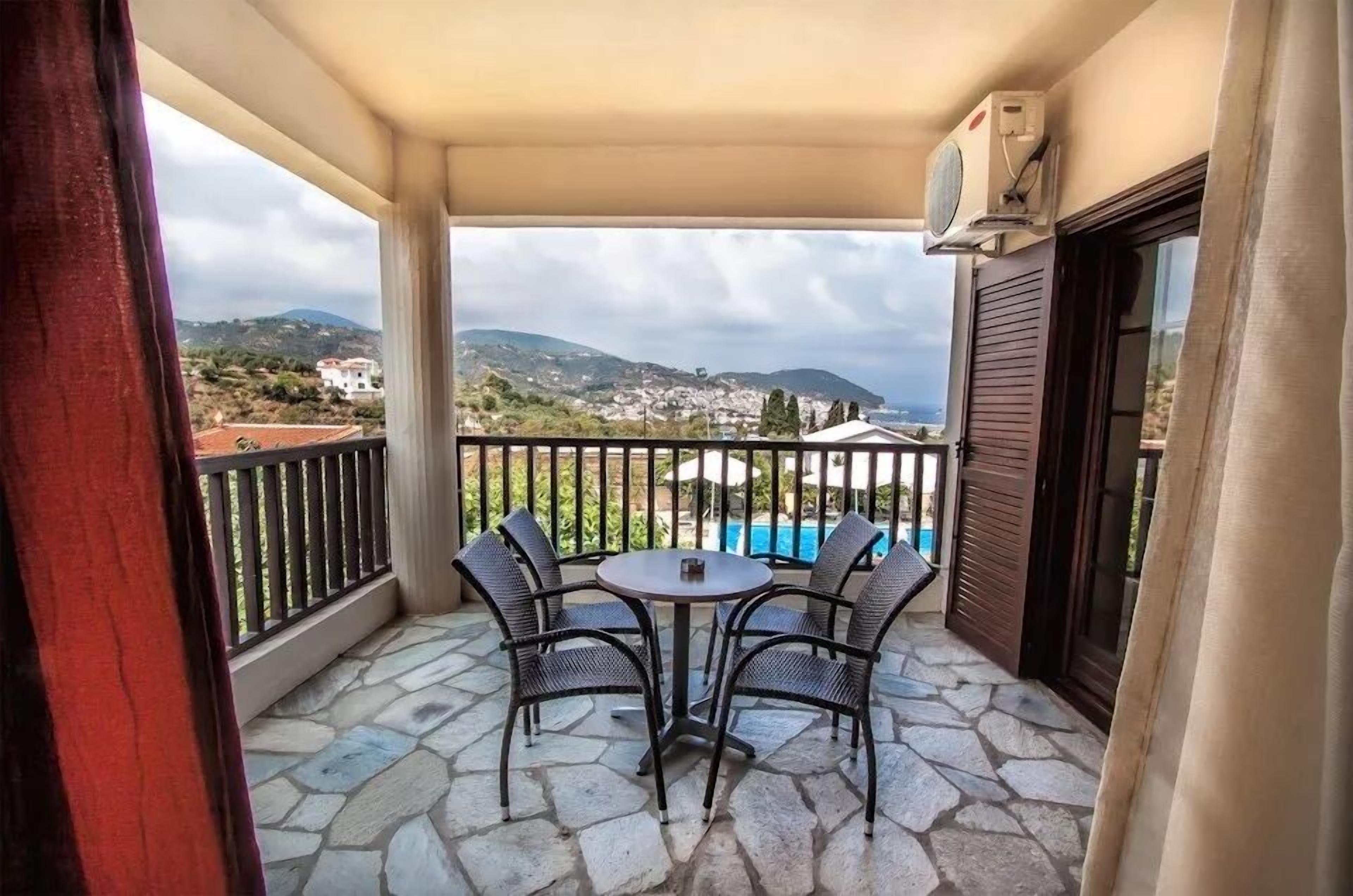 A balcony with a stone floor, a table with four chairs, and a view of a mountainous landscape.