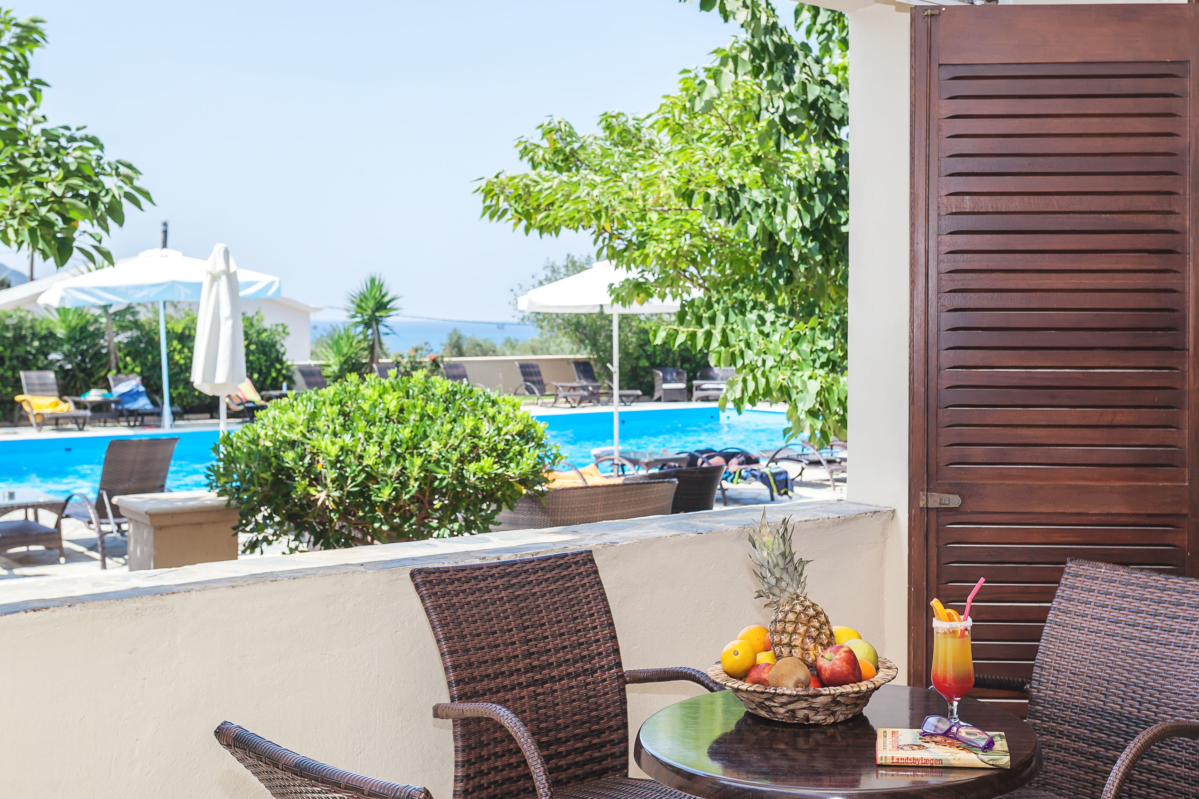 Een balkon met rieten stoelen, een tafel met fruit en een drankje, en uitzicht op een zwembad en parasols.