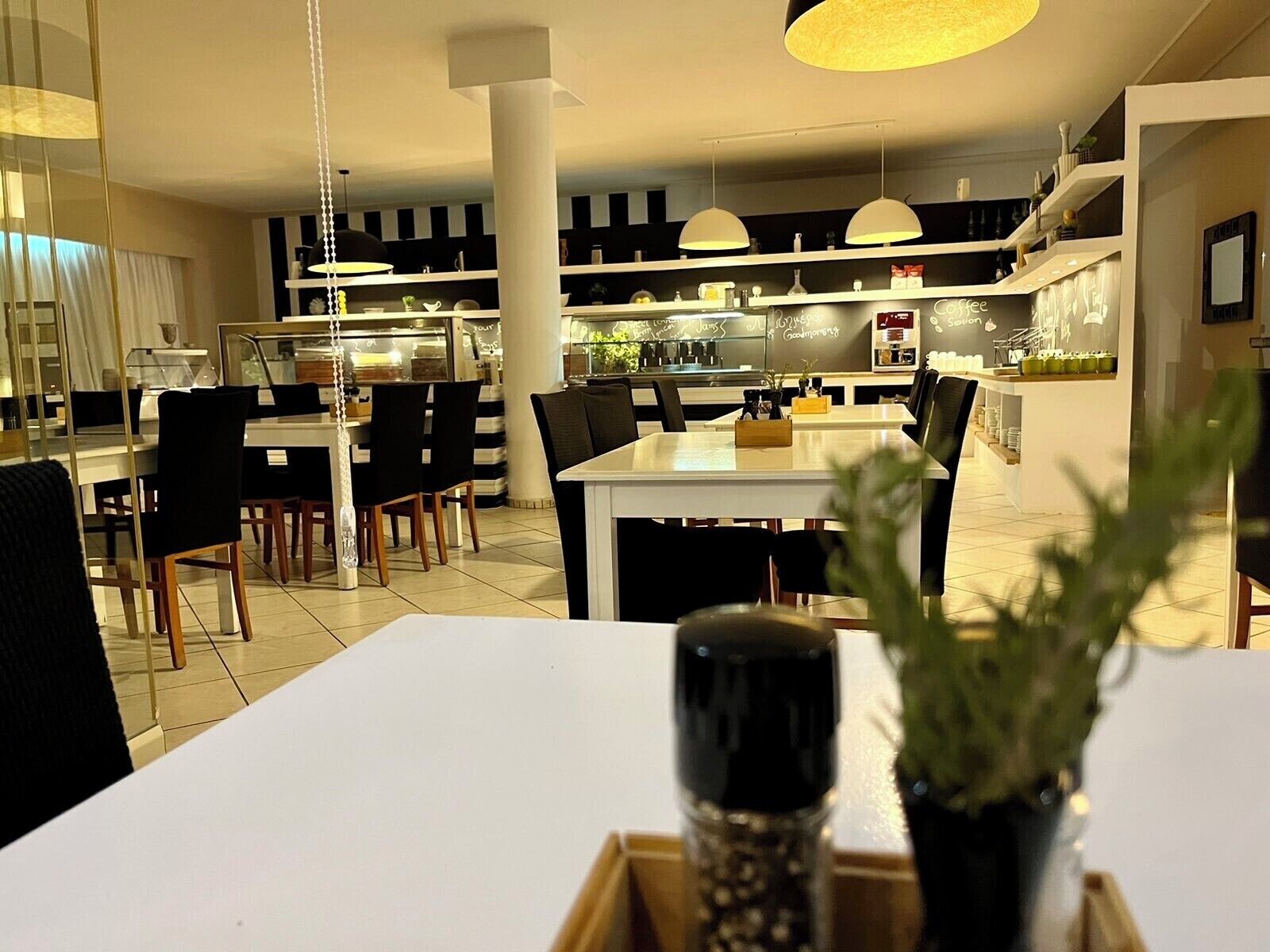Un espace de repas moderne avec des tables et des chaises blanches, un îlot central agrémenté d’une plante, et une cuisine en arrière-plan.