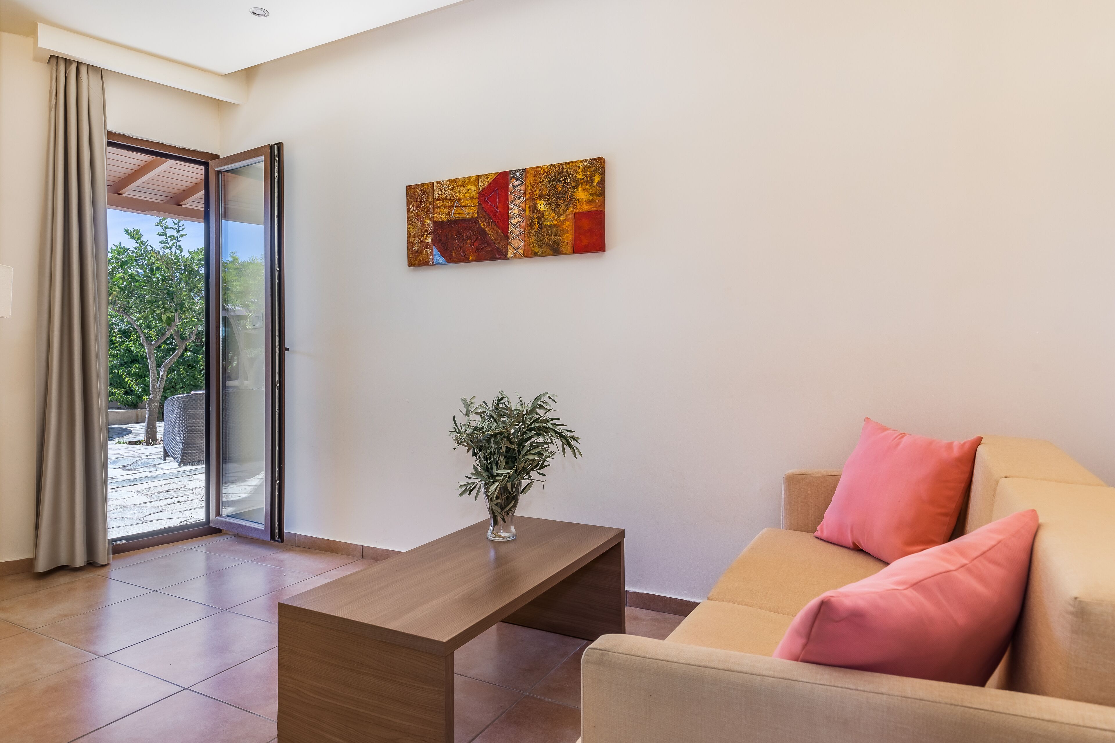 A modern living room with a sliding glass door leading to an outdoor area, a wooden coffee table, a beige sofa with pink cushions, and a piece of abstract art on the wall.