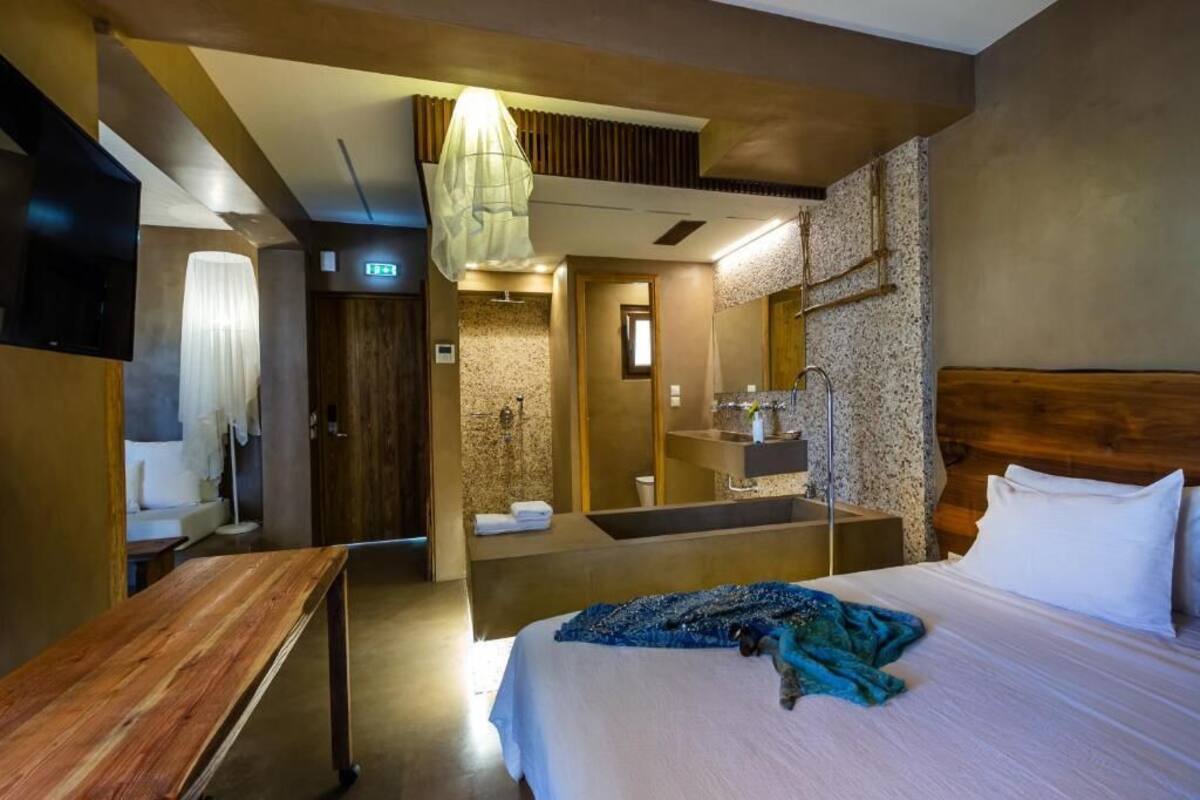 A hotel room with a bed, a wooden bench, a mirror, a TV, and a bathroom with a shower and sink.