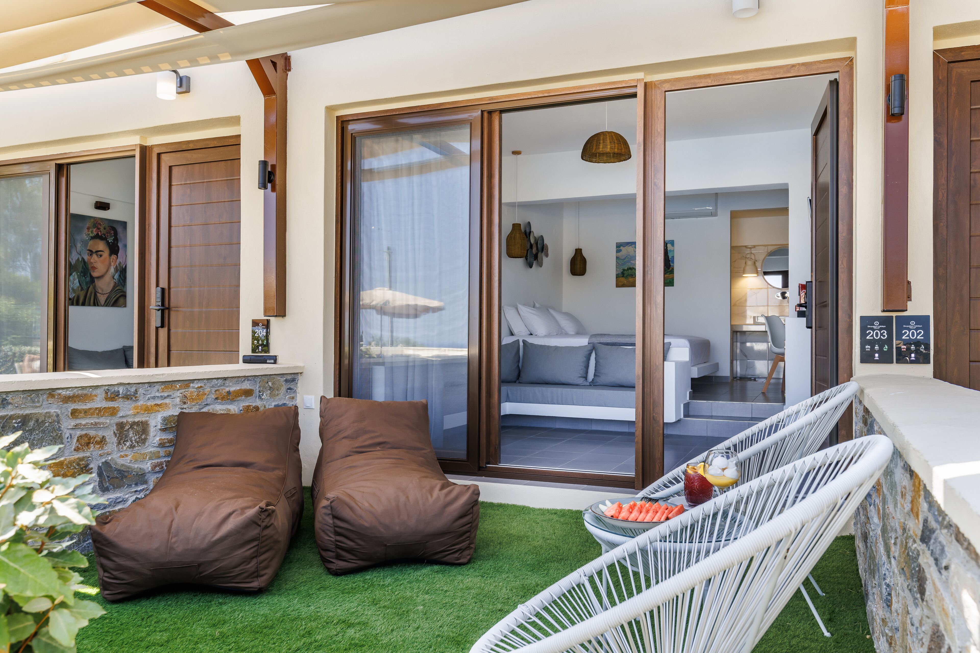Ein moderner Außenbereich mit Sitzgelegenheiten, darunter zwei braune Sitzsäcke und ein weißer Korbstuhl, ein kleiner Tisch mit Snacks und Blick in ein Schlafzimmer mit Bett und Sofa.