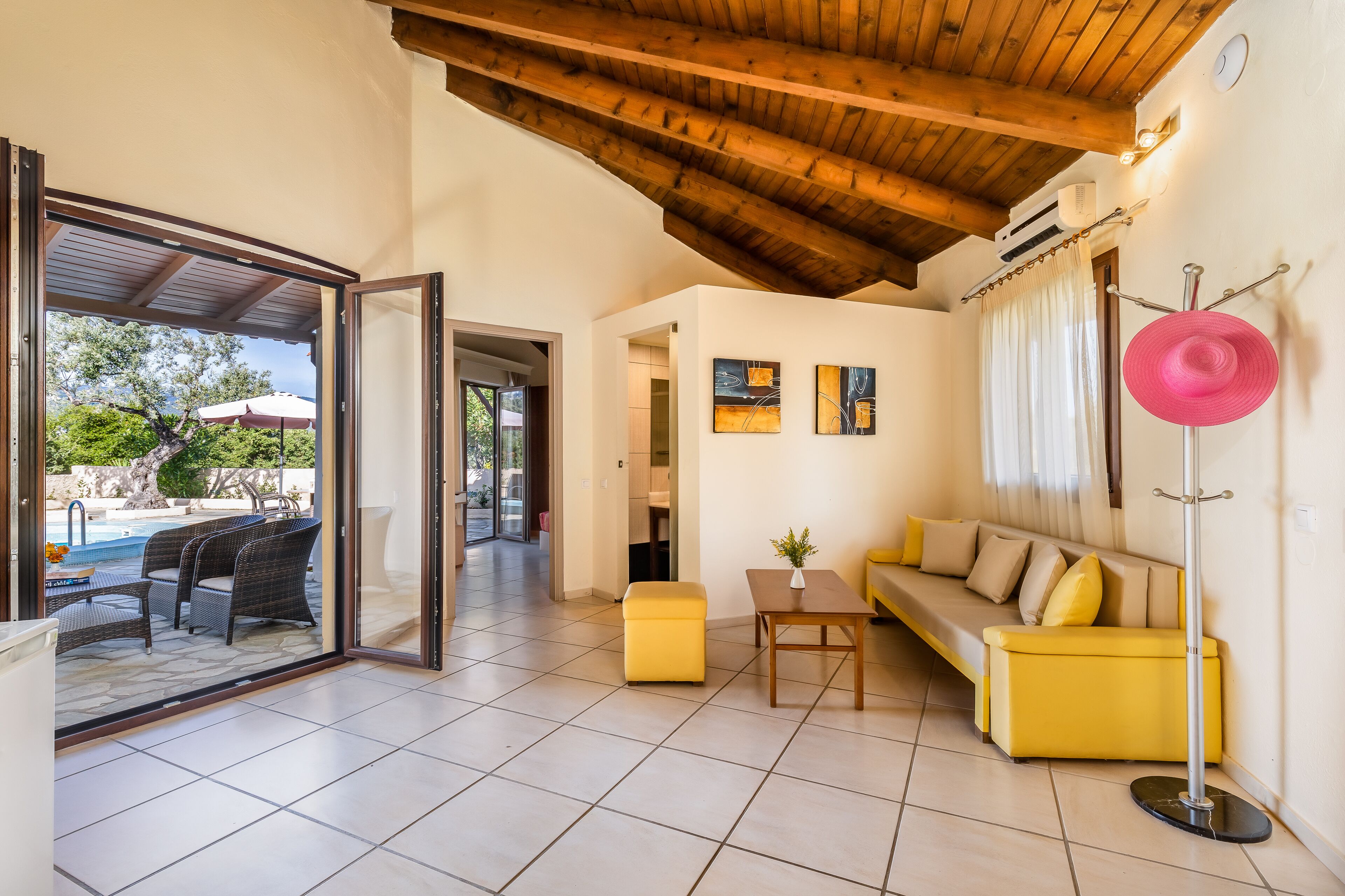 A modern living room with a yellow sofa, a small table, and a floor lamp. There is a glass door leading to an outdoor area with a pool and furniture.