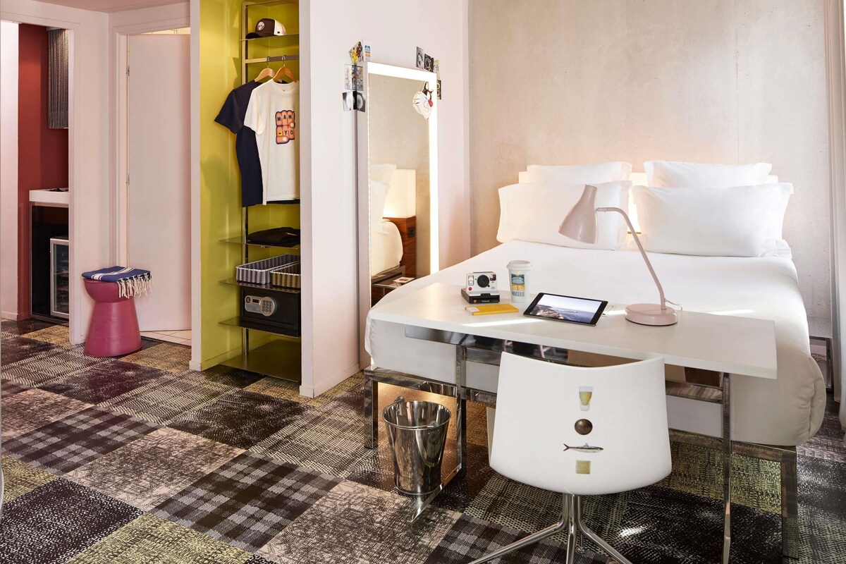 Een hotelkamer met een bed, nachtkastje, lamp, stoel en een klein bureau met een tablet en camera.