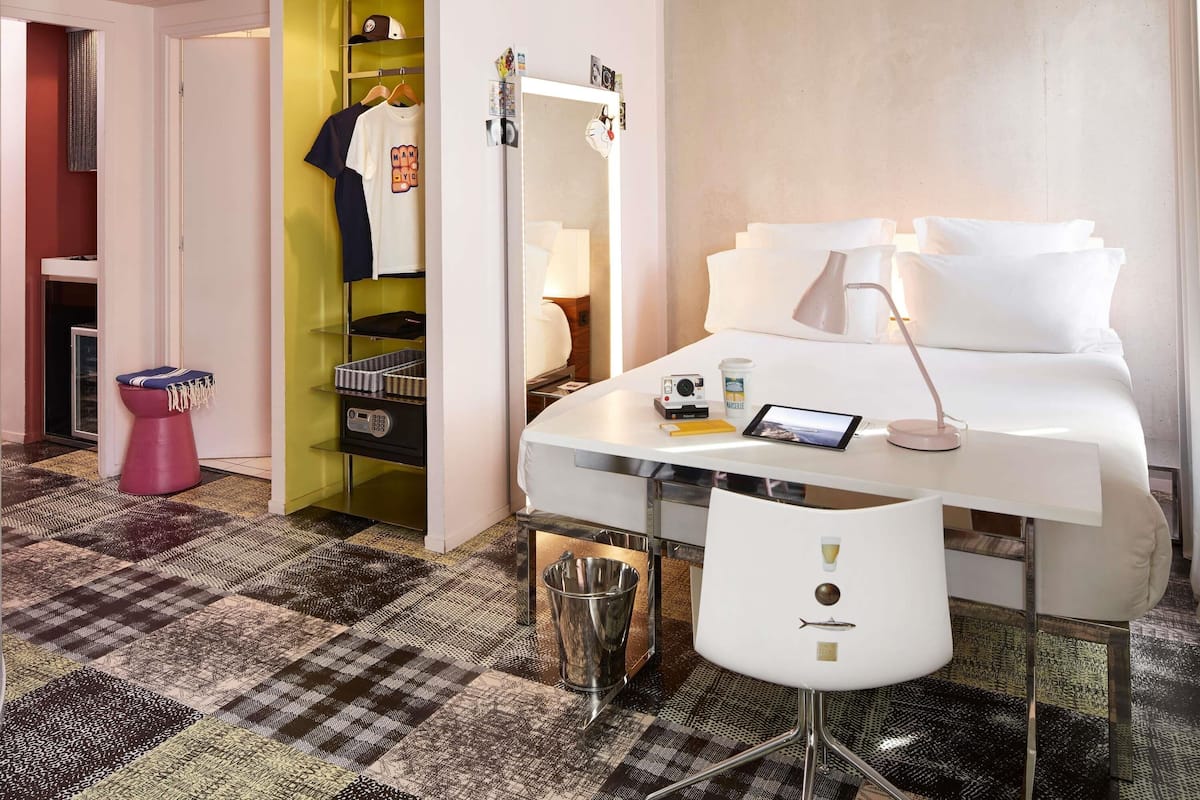 Une chambre d’hôtel comprenant un lit, une table de chevet, une lampe, une chaise et un petit bureau avec une tablette et un appareil photo.