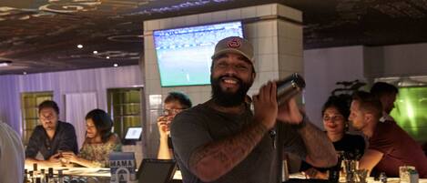 A bar area with a counter, bottles, and a television screen displaying a sports event.