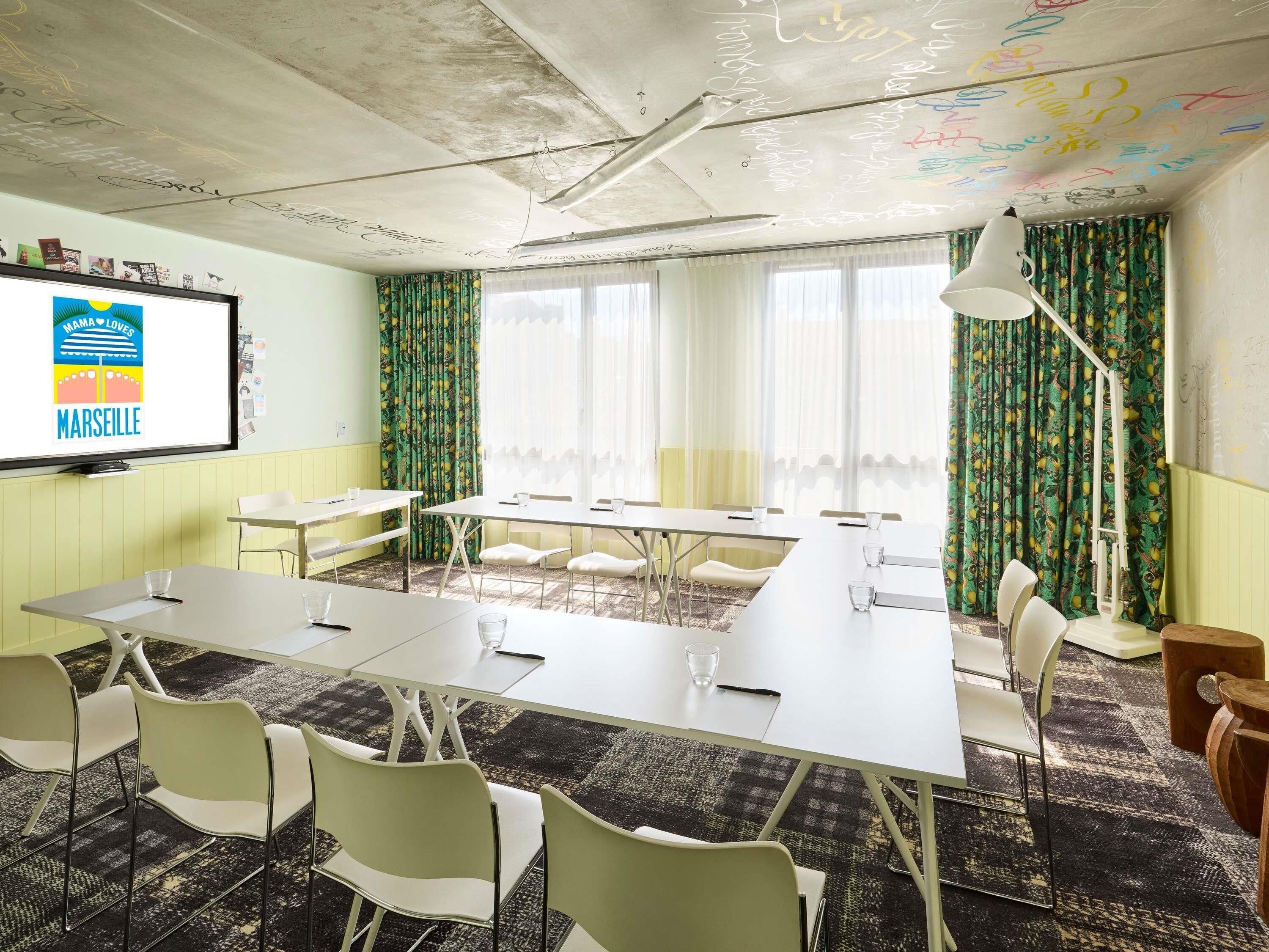 Uma sala de conferência com uma mesa comprida, cadeiras brancas, uma tela de projetor e cortinas estampadas.