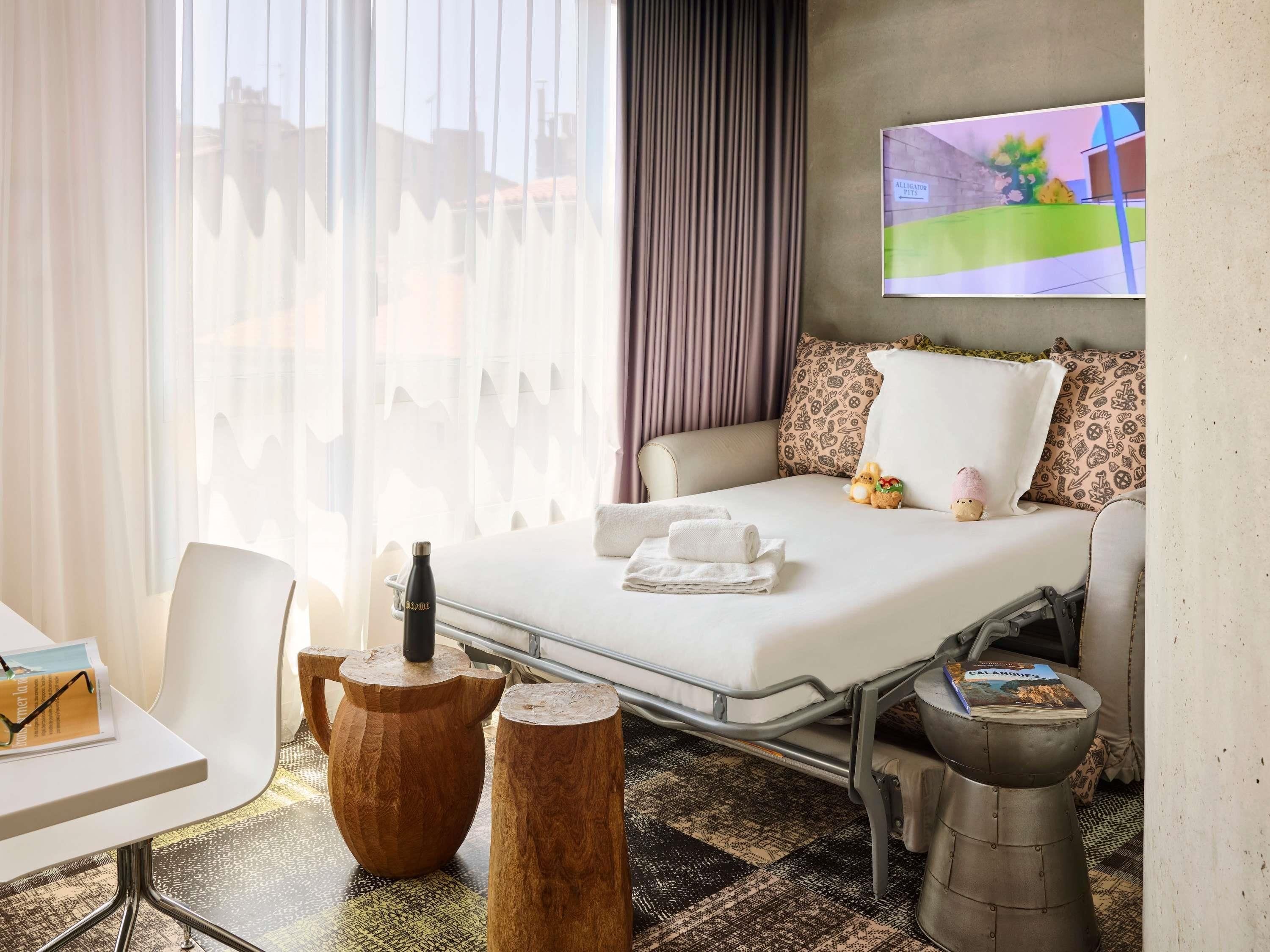 A hotel room with a bed, a TV, a table with a book, and a window with curtains.