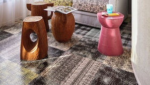 Un salon moderne avec un canapé, des coussins à motifs, une table basse et un tabouret rose original. Un panneau d’accueil encadré est accroché au mur.