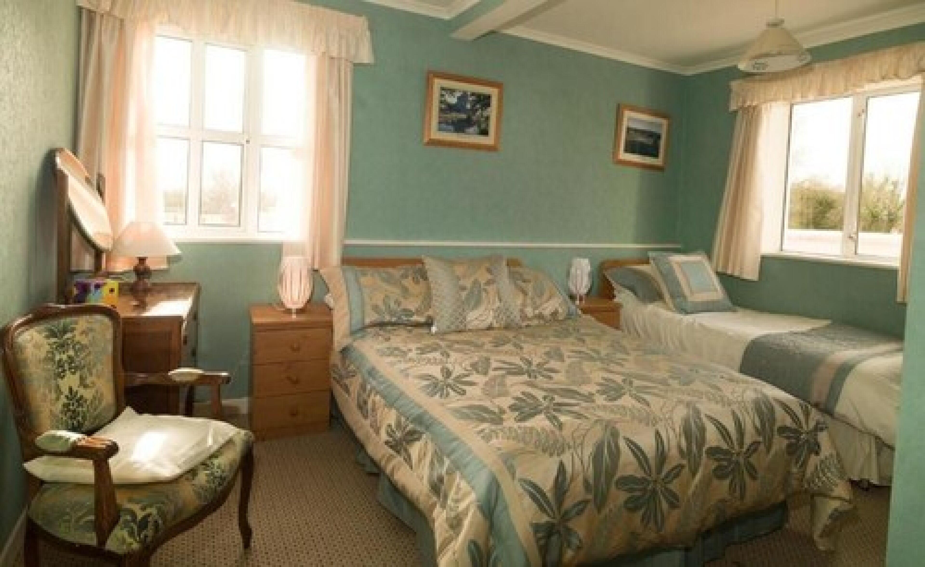 Ein Schlafzimmer mit einem Bett, zwei Nachttischen, einem Sessel und zwei mit Vorhängen versehenen Fenstern.