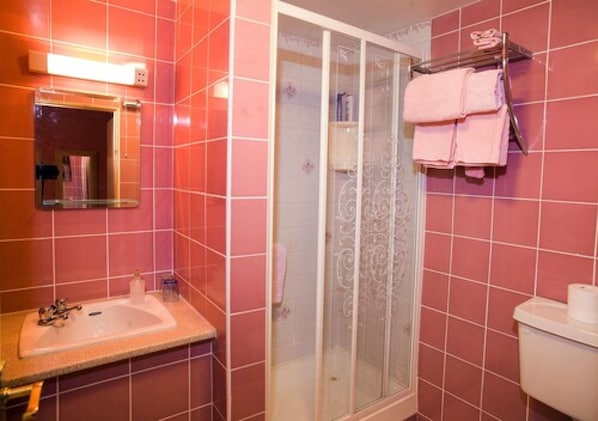Un baño con azulejos rosas, una ducha con mampara de vidrio, un lavamanos con espejo y un perchero con toallas.