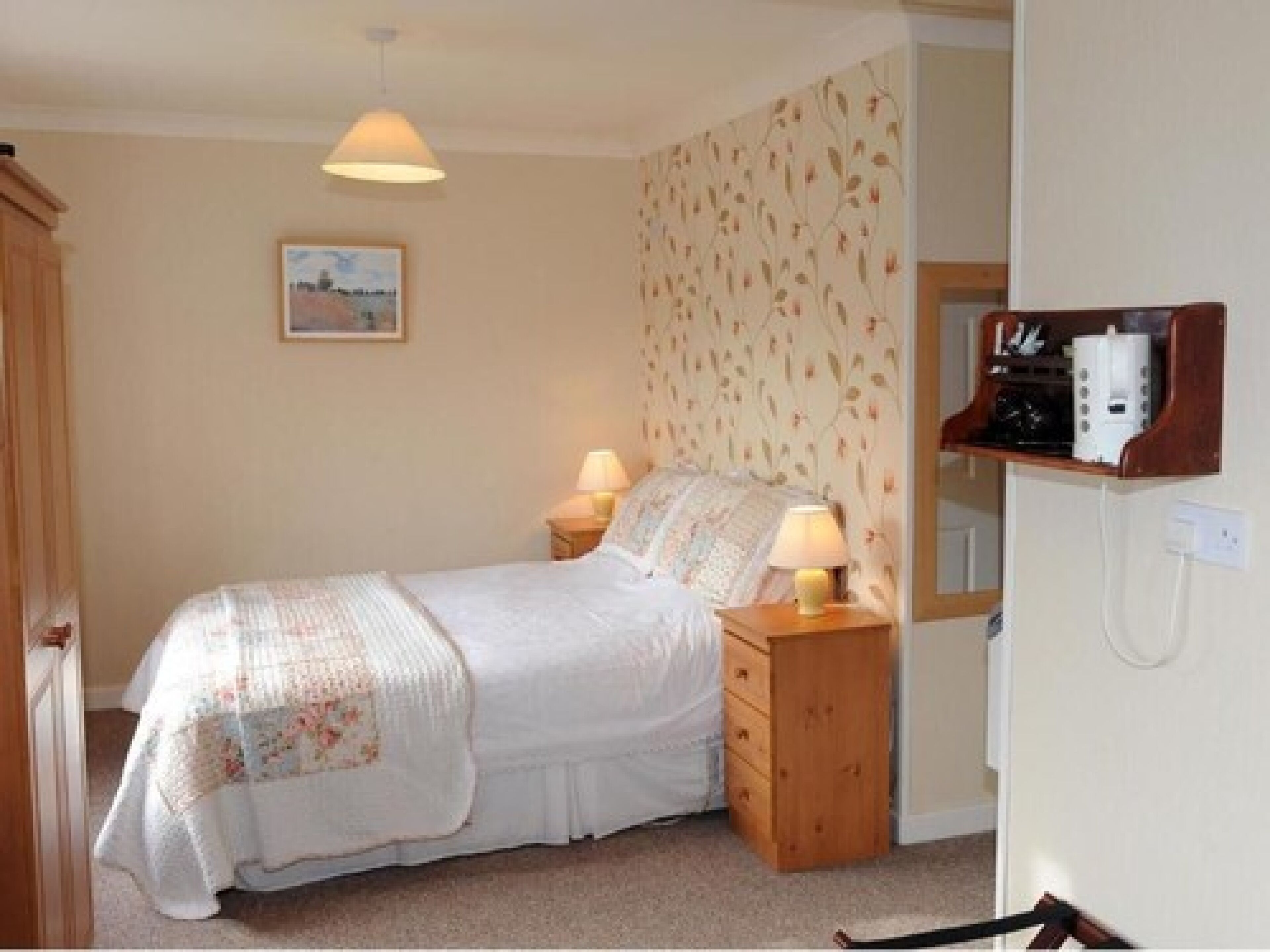 A bedroom with a bed, two bedside lamps, a wooden nightstand, a wall-mounted shelf with a radio, and a framed picture on the wall.