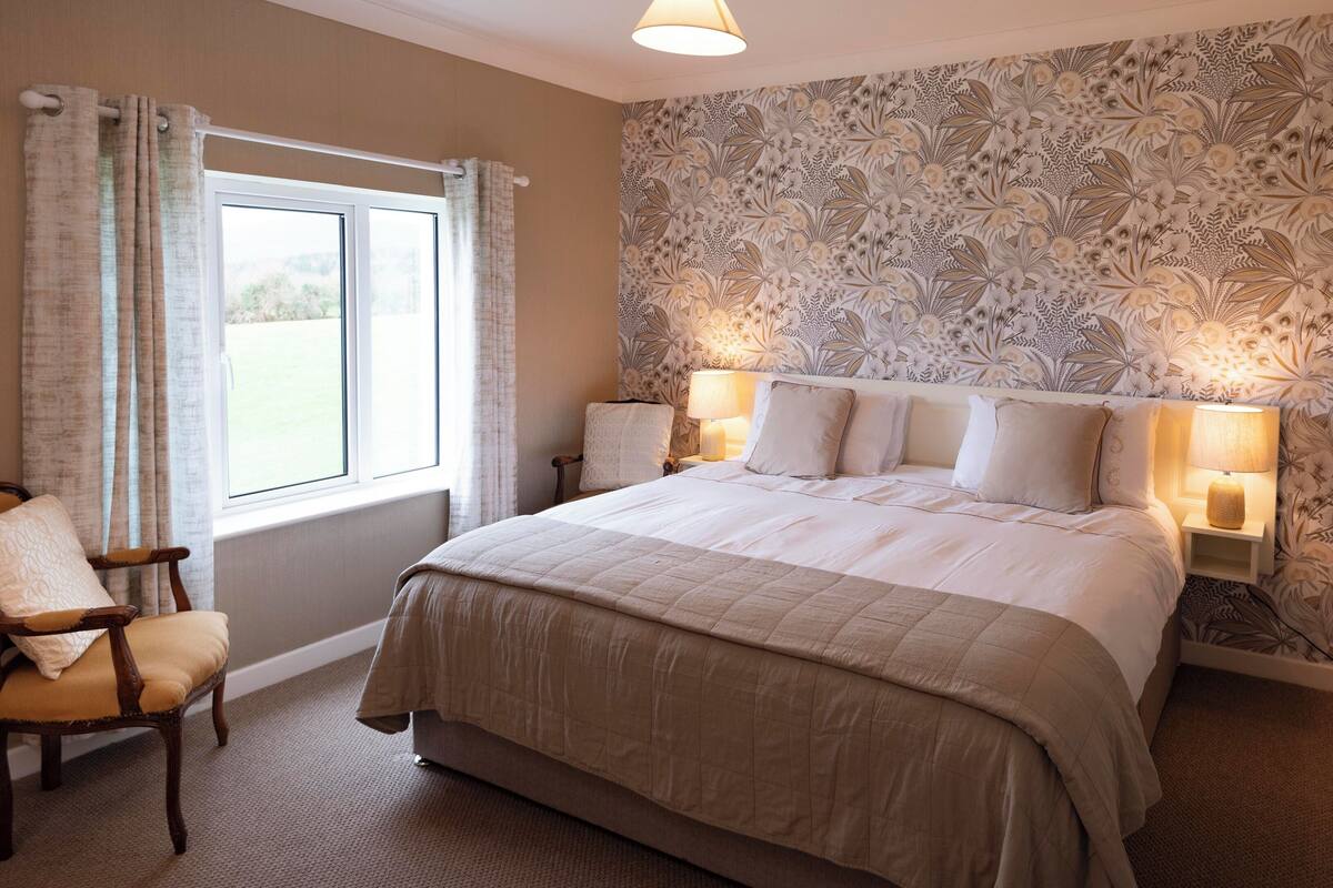 A bedroom with a large bed, two bedside lamps, a chair, and a window with curtains.