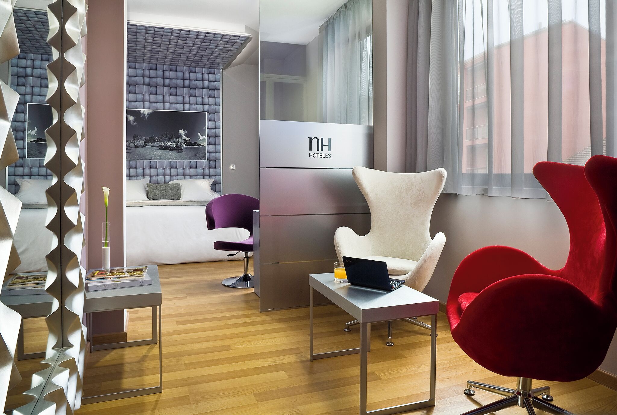 Una habitación de hotel moderna con sofá, sillas rojas, una pequeña mesa con una laptop, un vaso de jugo de naranja y un espejo de diseño único.