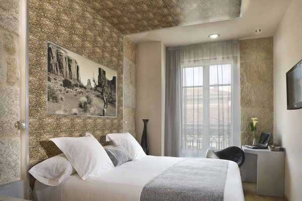 Un dormitorio con cama, escritorio, silla, ventana con cortinas y televisor montado en la pared.