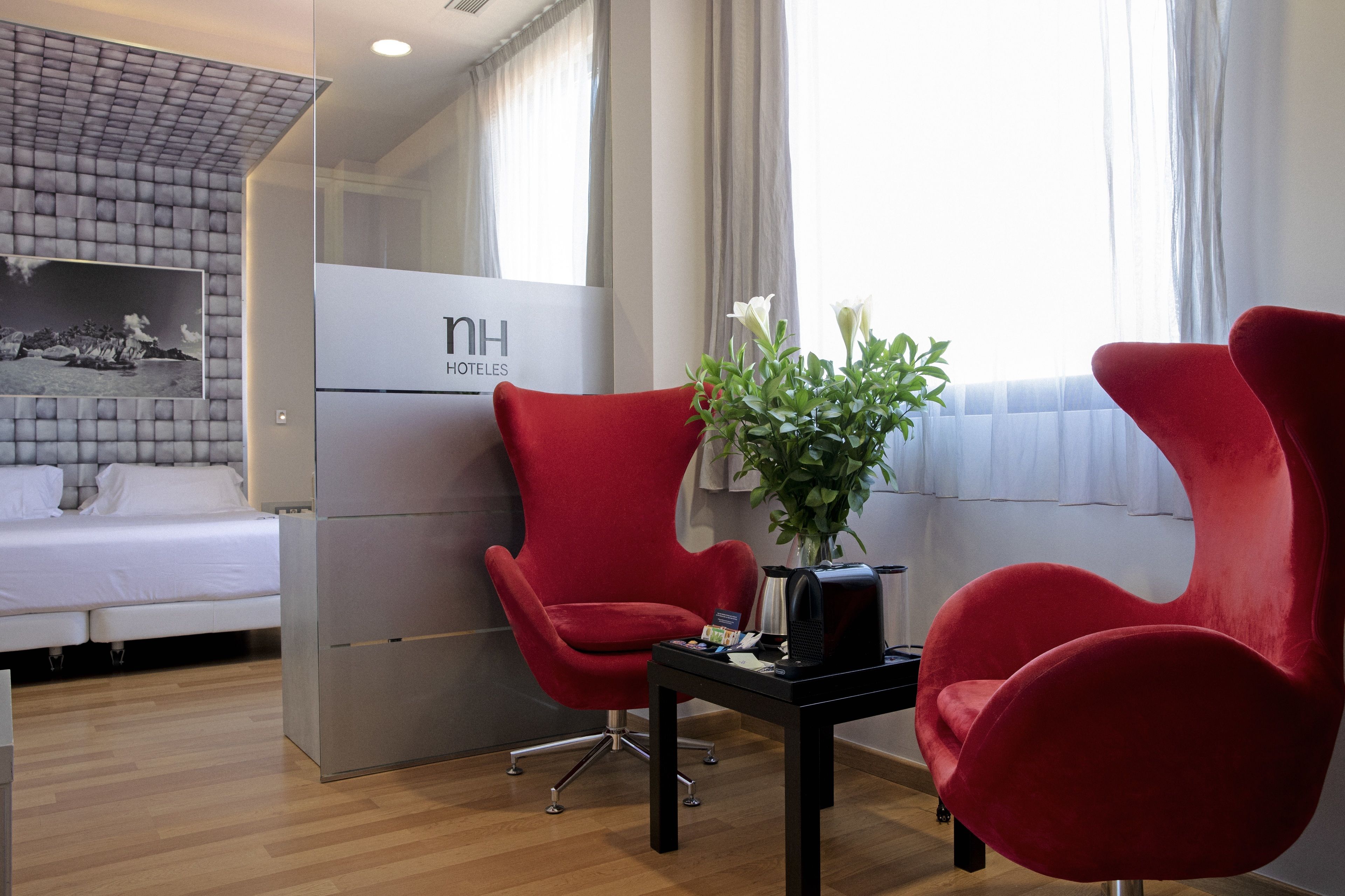 Una habitación de hotel con una cama, dos sillones rojos, una mesita con un jarrón de flores y un cartel que dice "nh hotels".