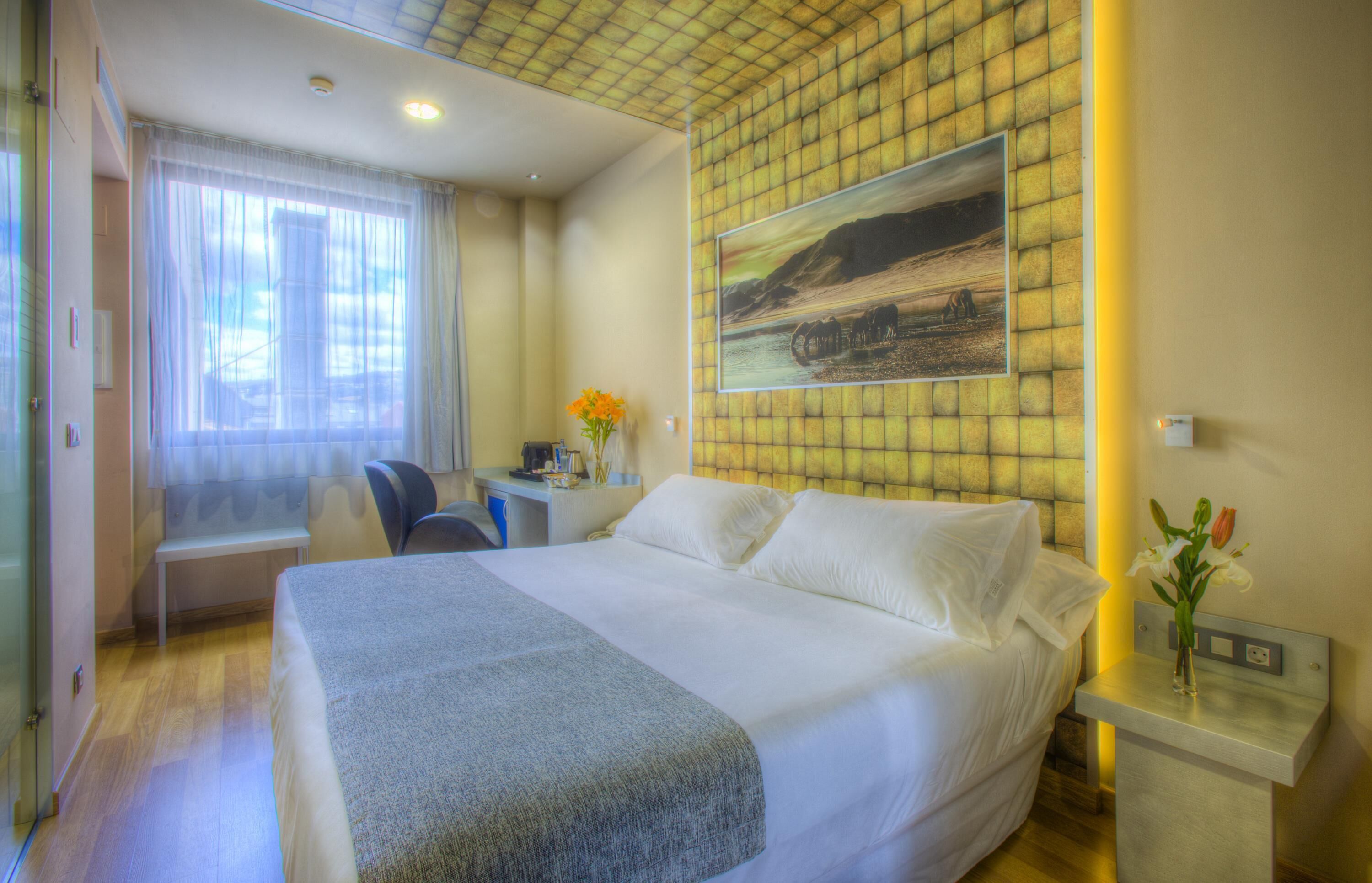 Une chambre d’hôtel avec un grand lit, un bureau, une chaise, une fenêtre avec des rideaux, un tableau au mur et un vase de fleurs sur la table de chevet.
