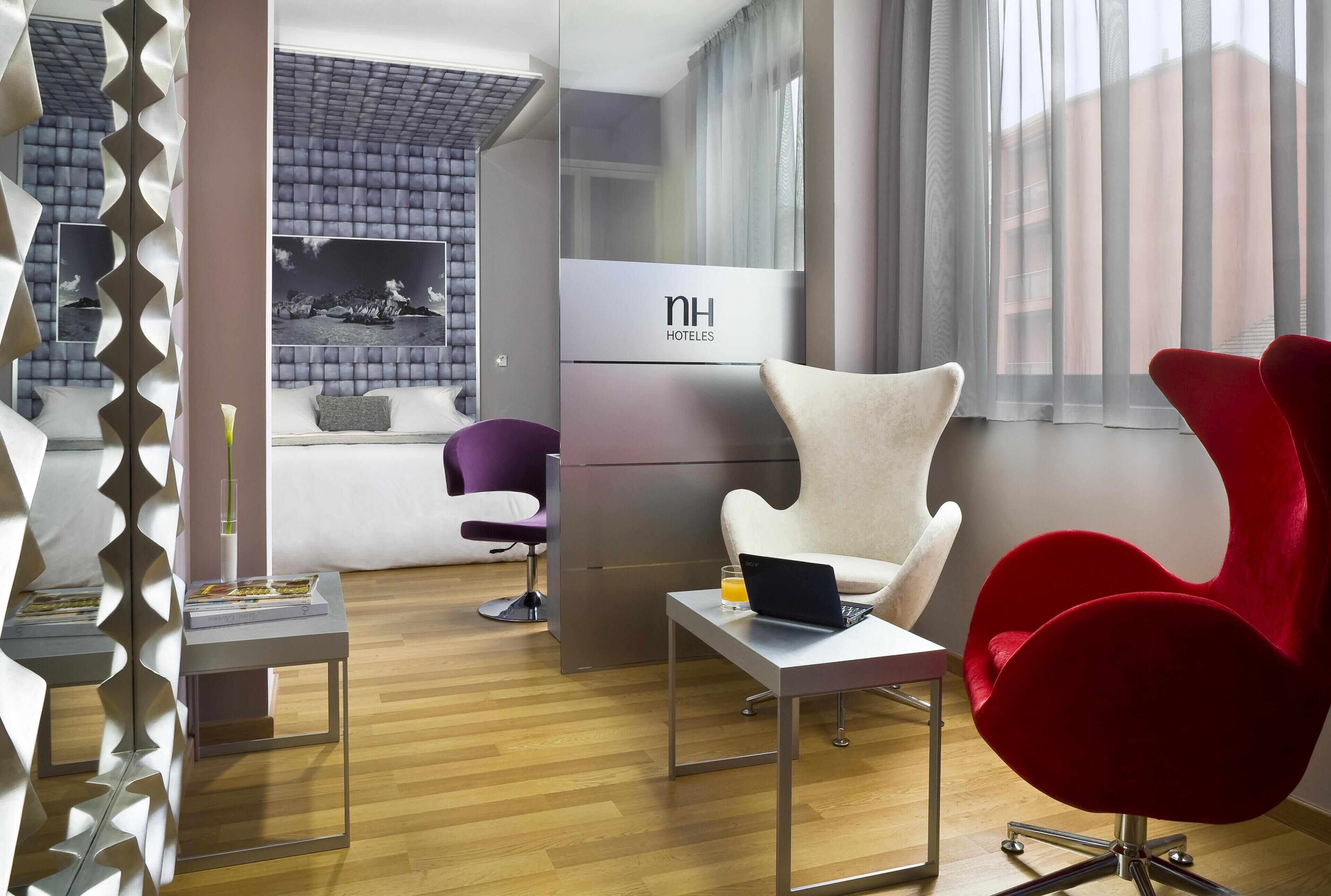 Una habitación de hotel moderna con una cama, un escritorio con una computadora portátil, una silla roja, una silla morada y un amplio ventanal con cortinas.