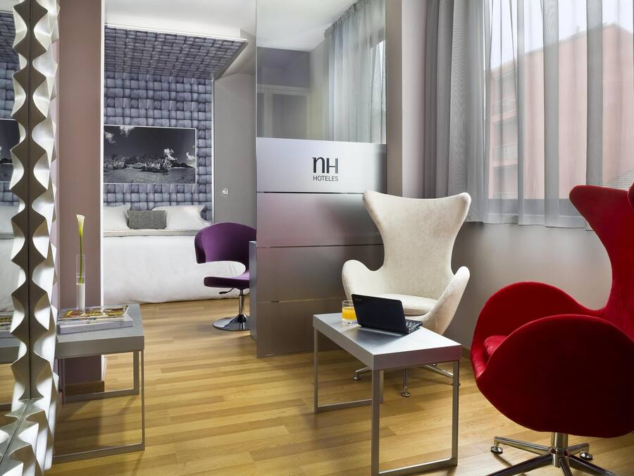 Una habitación de hotel moderna con una cama, un escritorio con una computadora portátil, una silla roja, una silla morada y un amplio ventanal con cortinas.