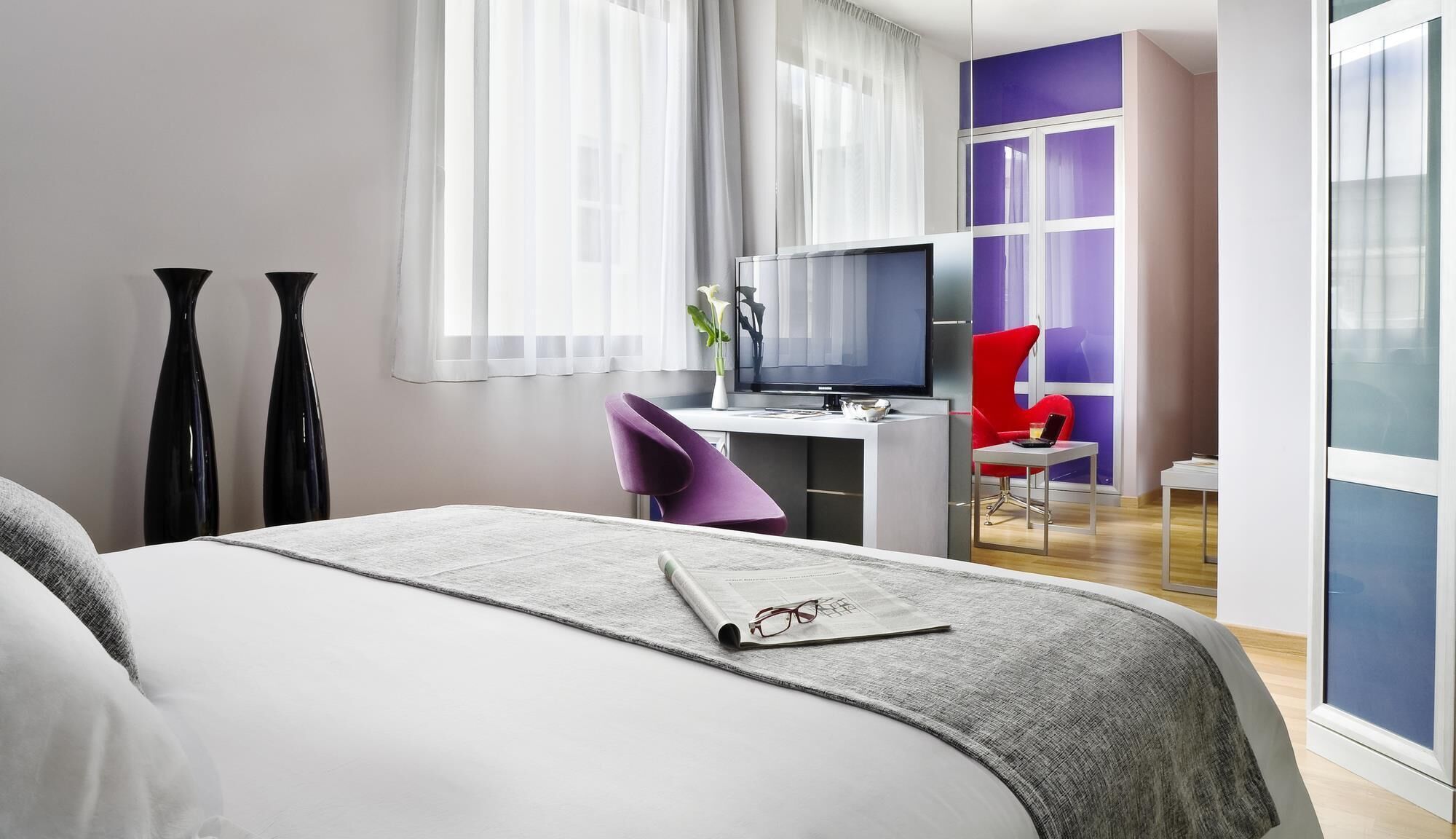 Una habitación de hotel moderna con una cama grande, un escritorio con computadora, una silla roja y un televisor.