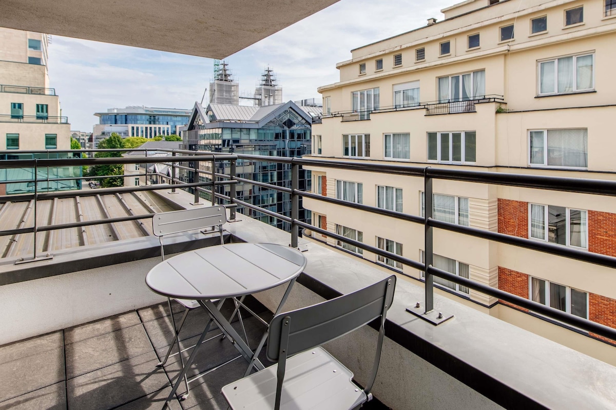 Un balcón con mesa y sillas, con vista a un paisaje urbano que incluye edificios y una obra en construcción.