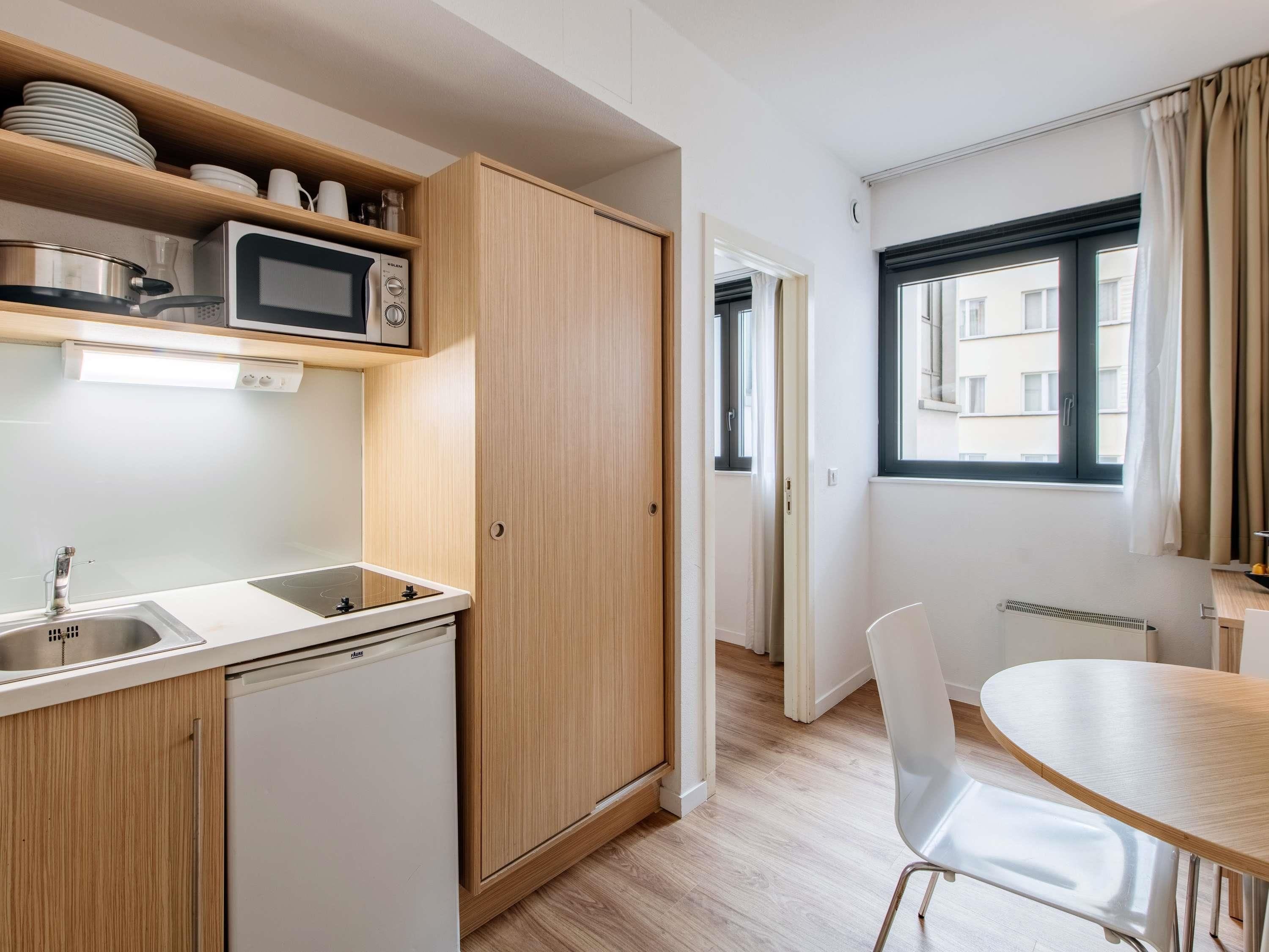Een compacte keuken met een magnetron, spoelbak en kastjes. Er is een eethoek met een tafel en stoelen.