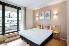 A hotel room with a large bed, a balcony with a table and chairs, and two wall-mounted lights. - Aparthotel Adagio Access Brussels Europe (Brussels)