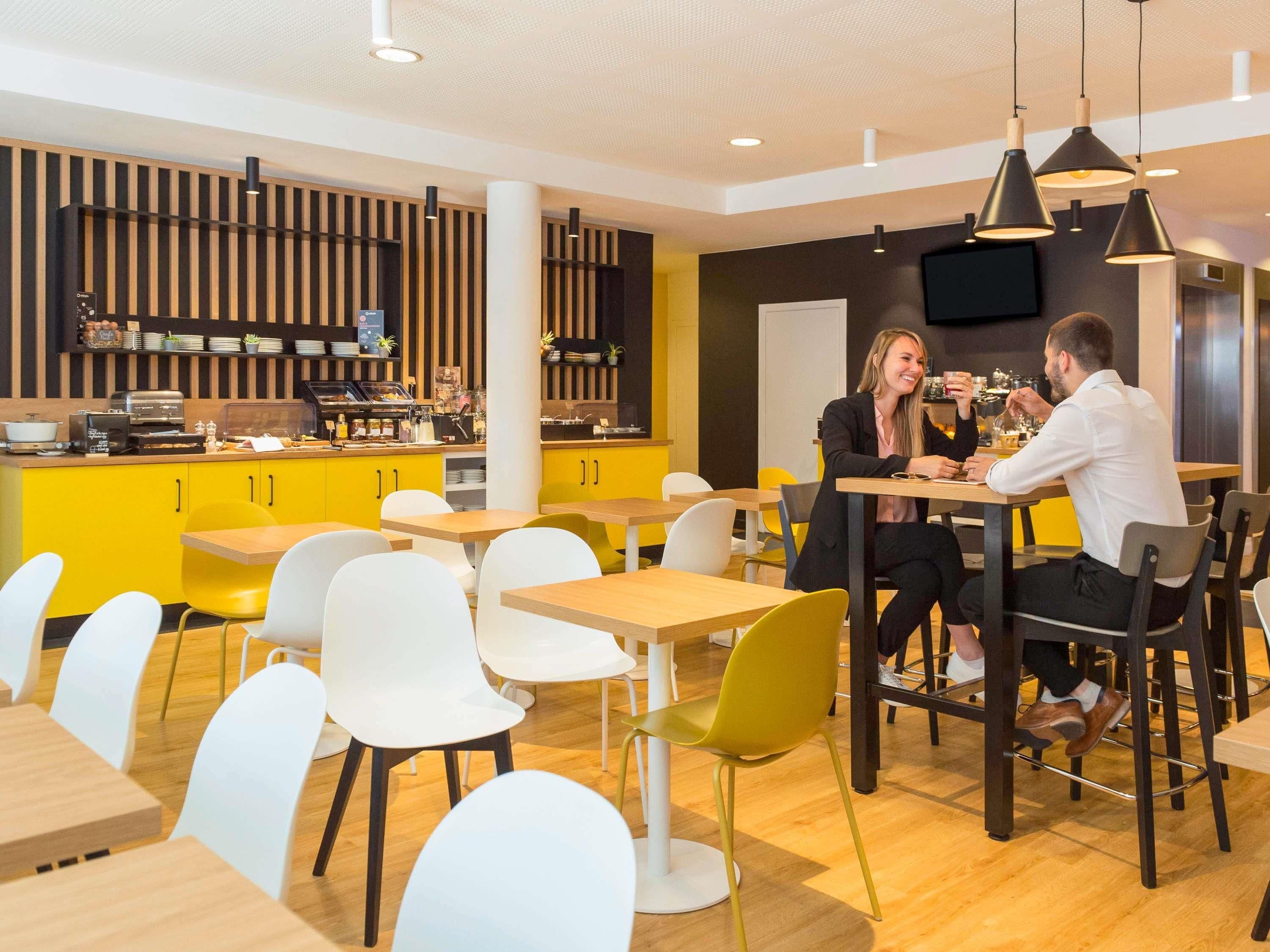 A modern dining area with yellow and white chairs, wooden tables, and a kitchen counter with appliances.