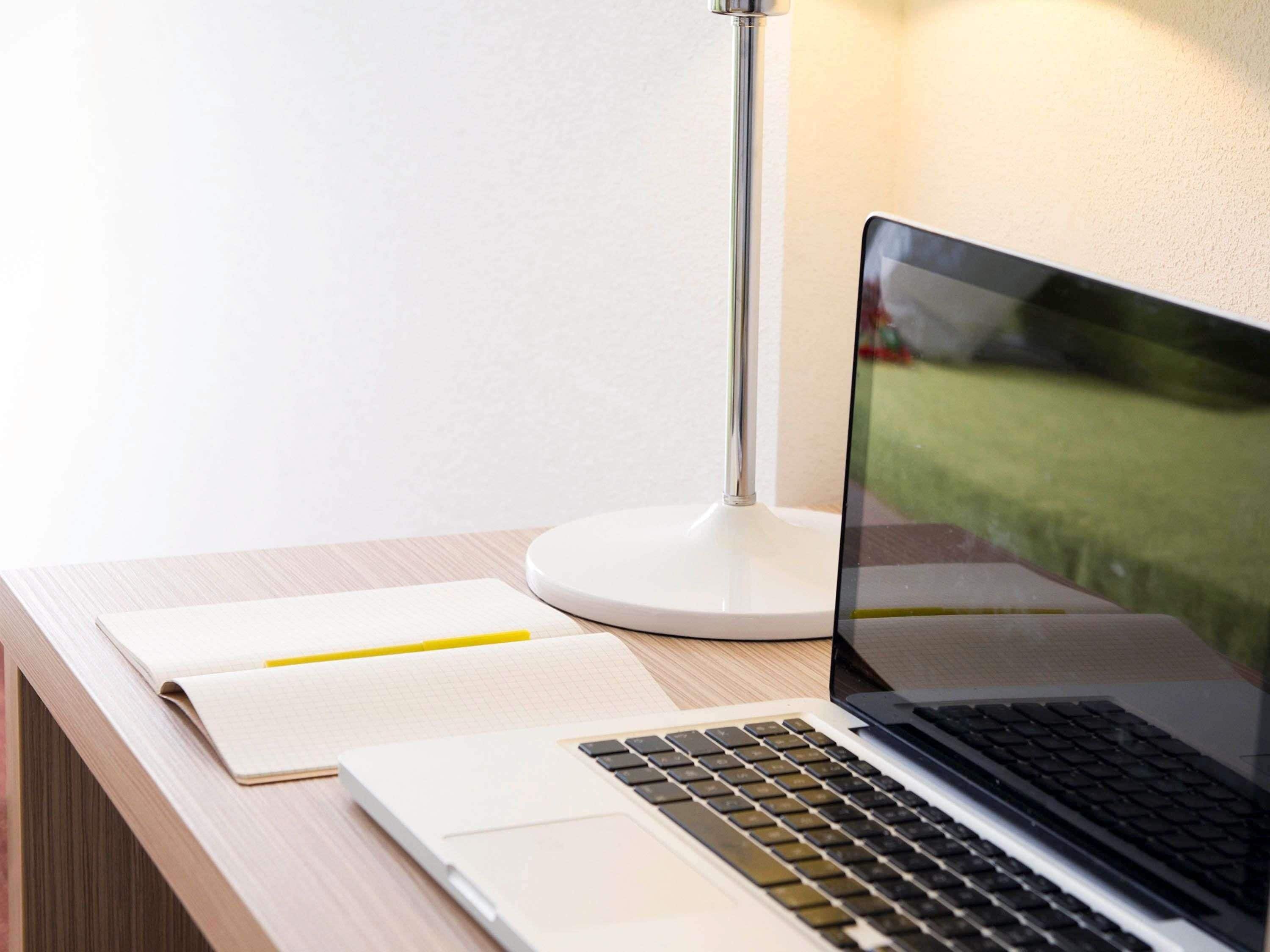 A laptop on a desk with a notebook and a pen, a lamp, and a window with a view of a green area.