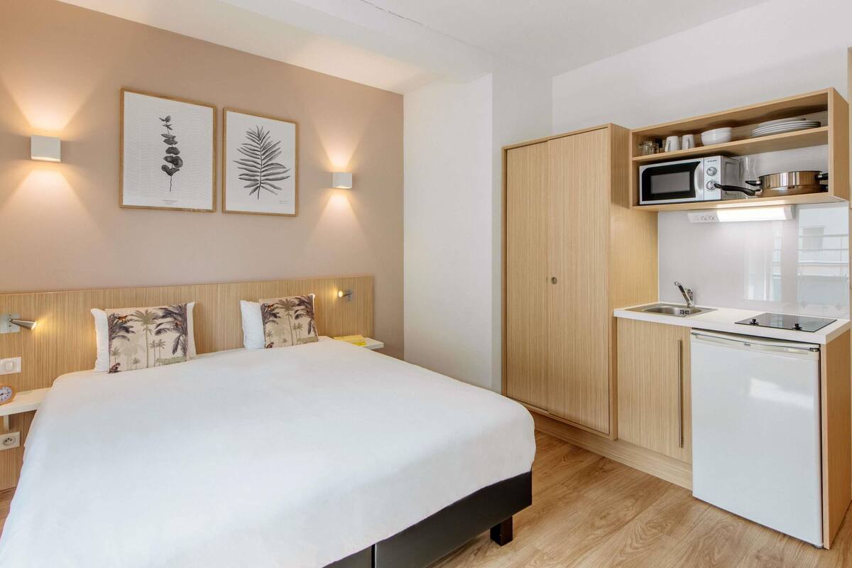 A compact hotel room with a bed, a small kitchenette, and a wall-mounted TV.