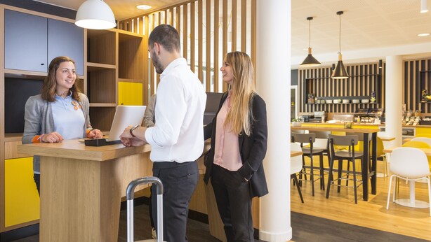 Un hall d’hôtel moderne comprenant un comptoir d’accueil, un espace salon avec des tables et des chaises, et un bar.