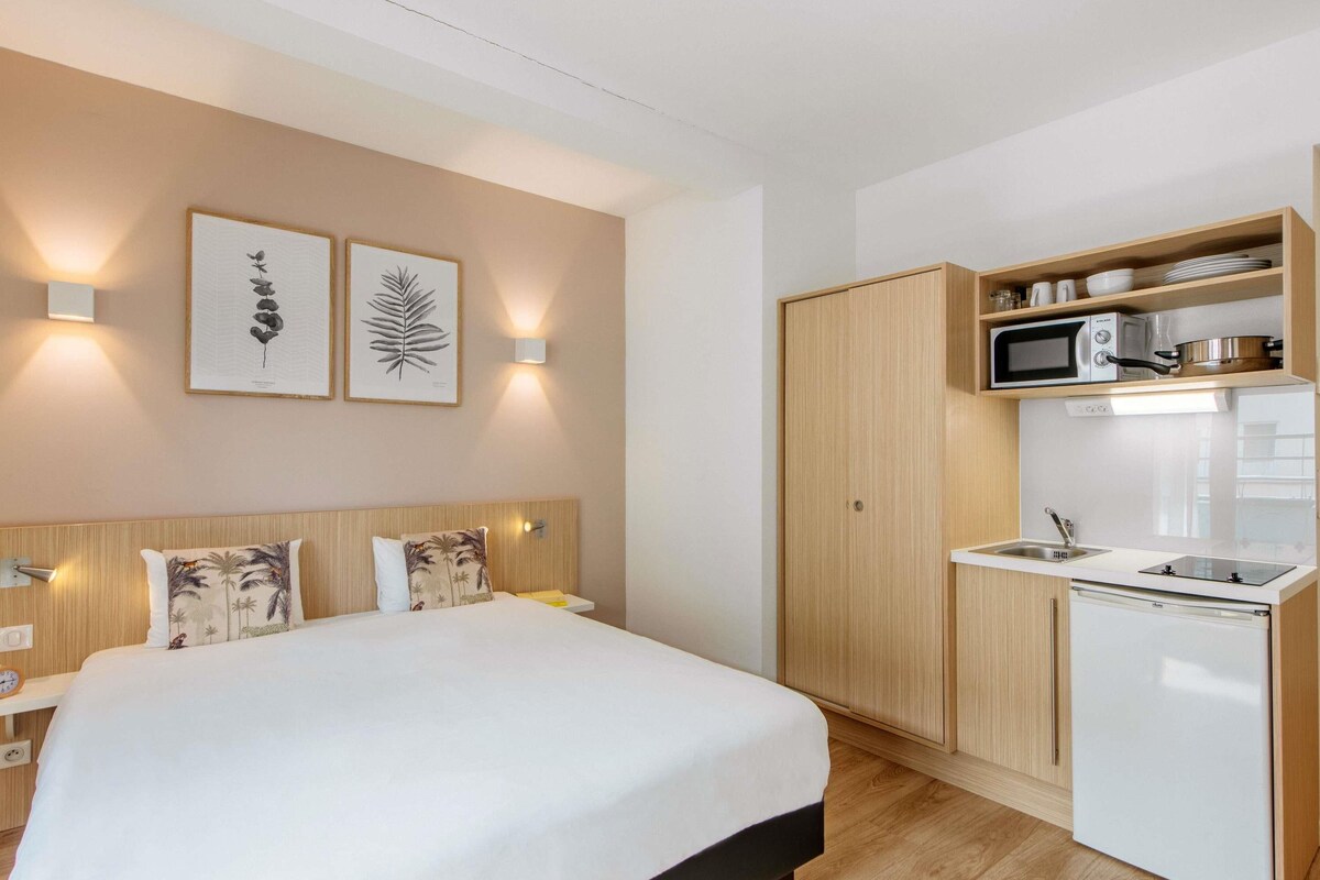 Una habitación de hotel compacta con cama, kitchenette pequeña y televisor montado en la pared.